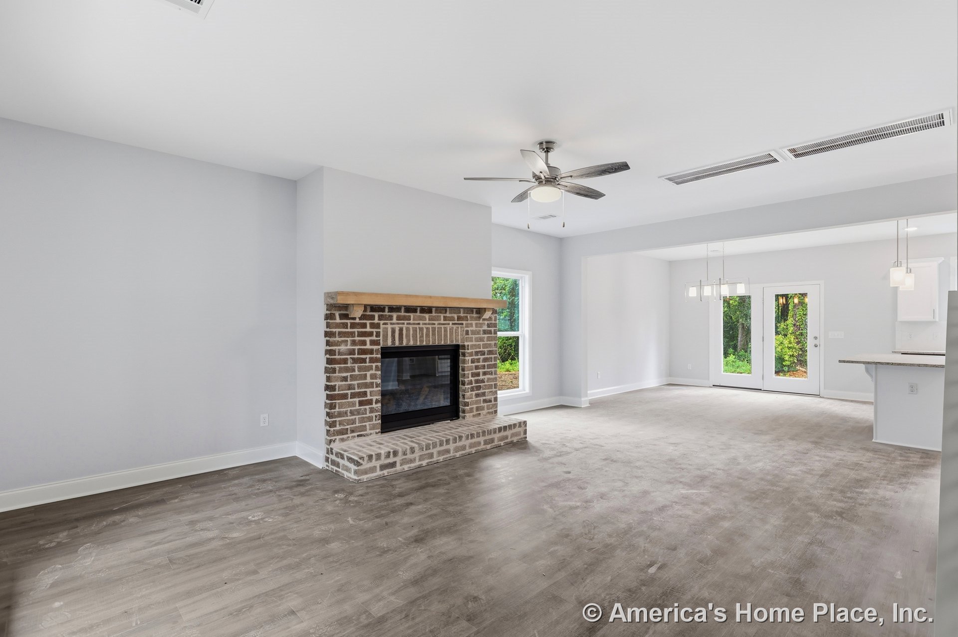 Brick fireplace with wood mantel anchors the living area, featuring hardwood flooring, ceiling fan with light fixture, large windows, and double glass doors leading outside; open