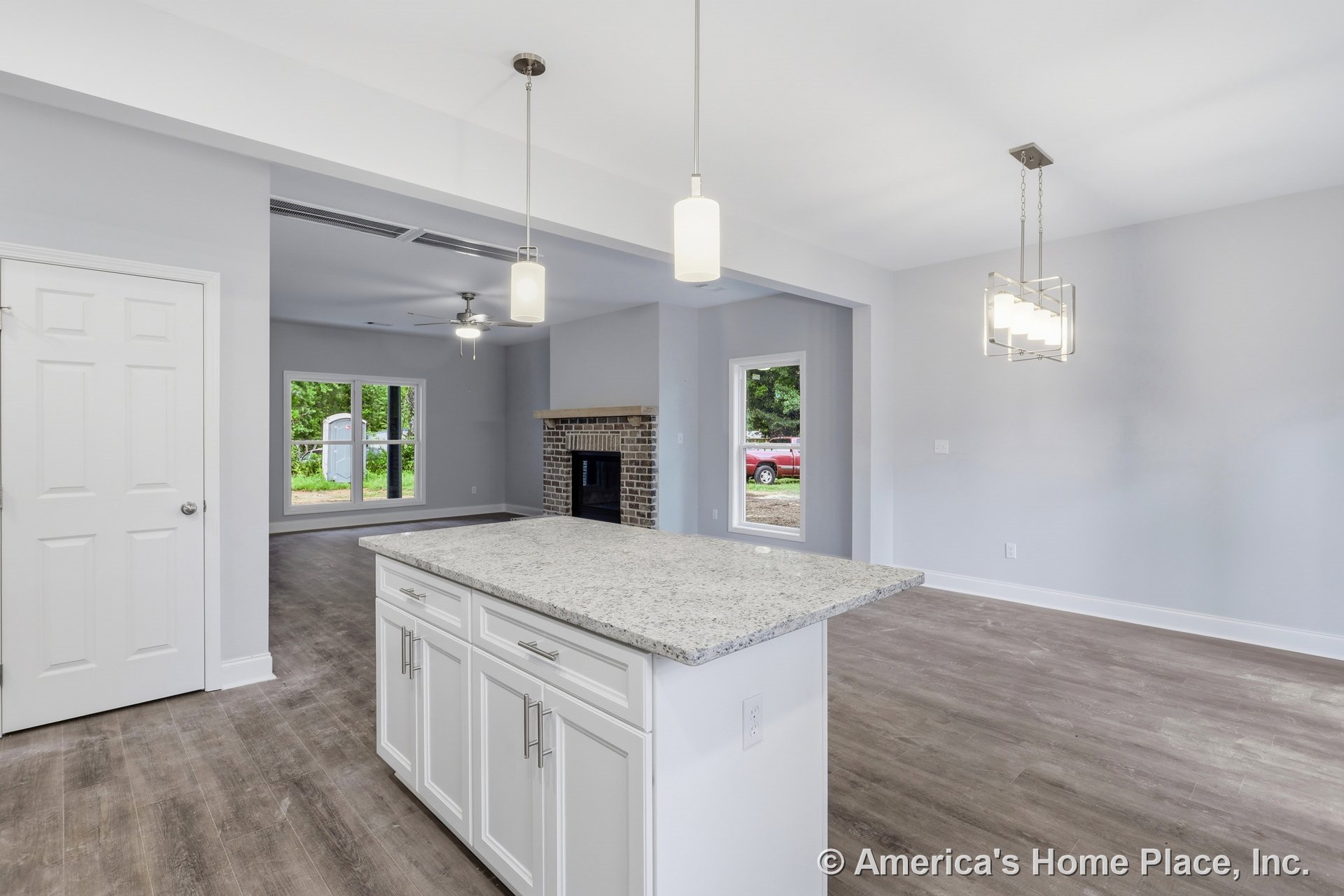 Open concept kitchen with white shaker cabinets, granite island, modern pendant lights, wood-look flooring, and large windows, overlooking living room with brick fireplace.