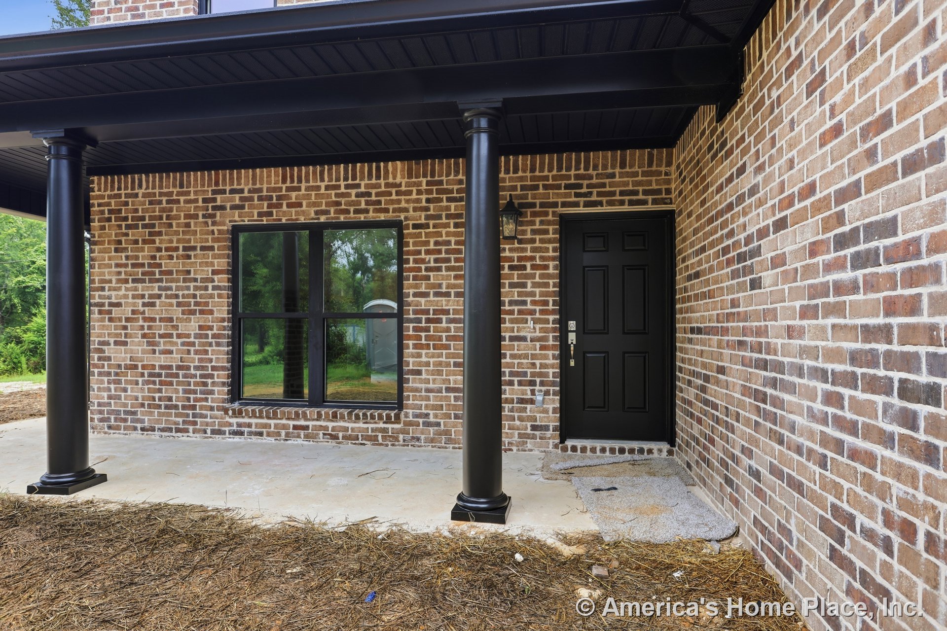Covered front porch with brick exterior walls, black columns, black framed window, black entry door, exterior lighting fixture, and black trim accents.