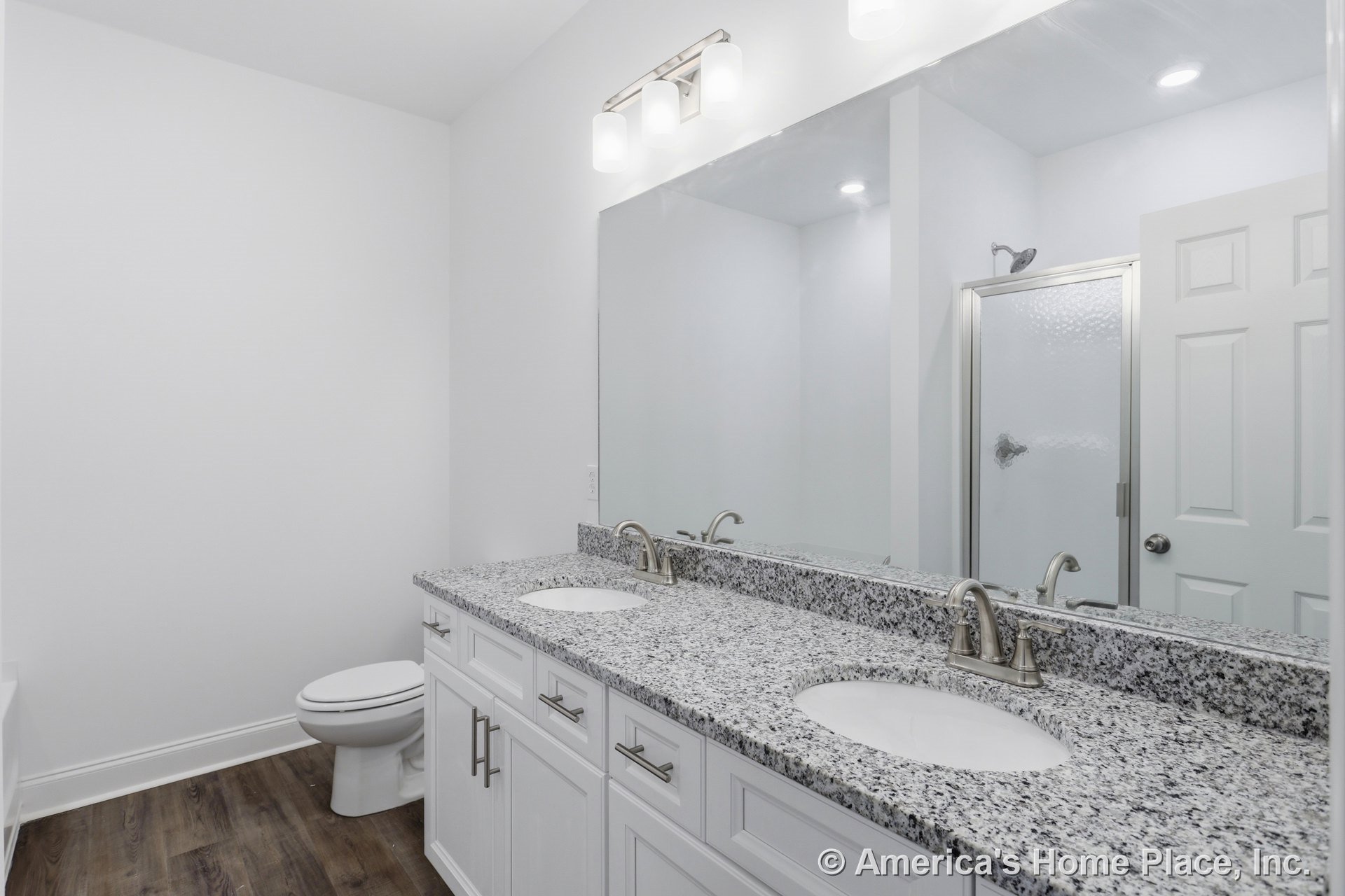 Double vanity with granite countertops and white shaker cabinets, large wall mirror, built-in overhead lighting, wood-look flooring, glass shower enclosure, and chrome faucets.