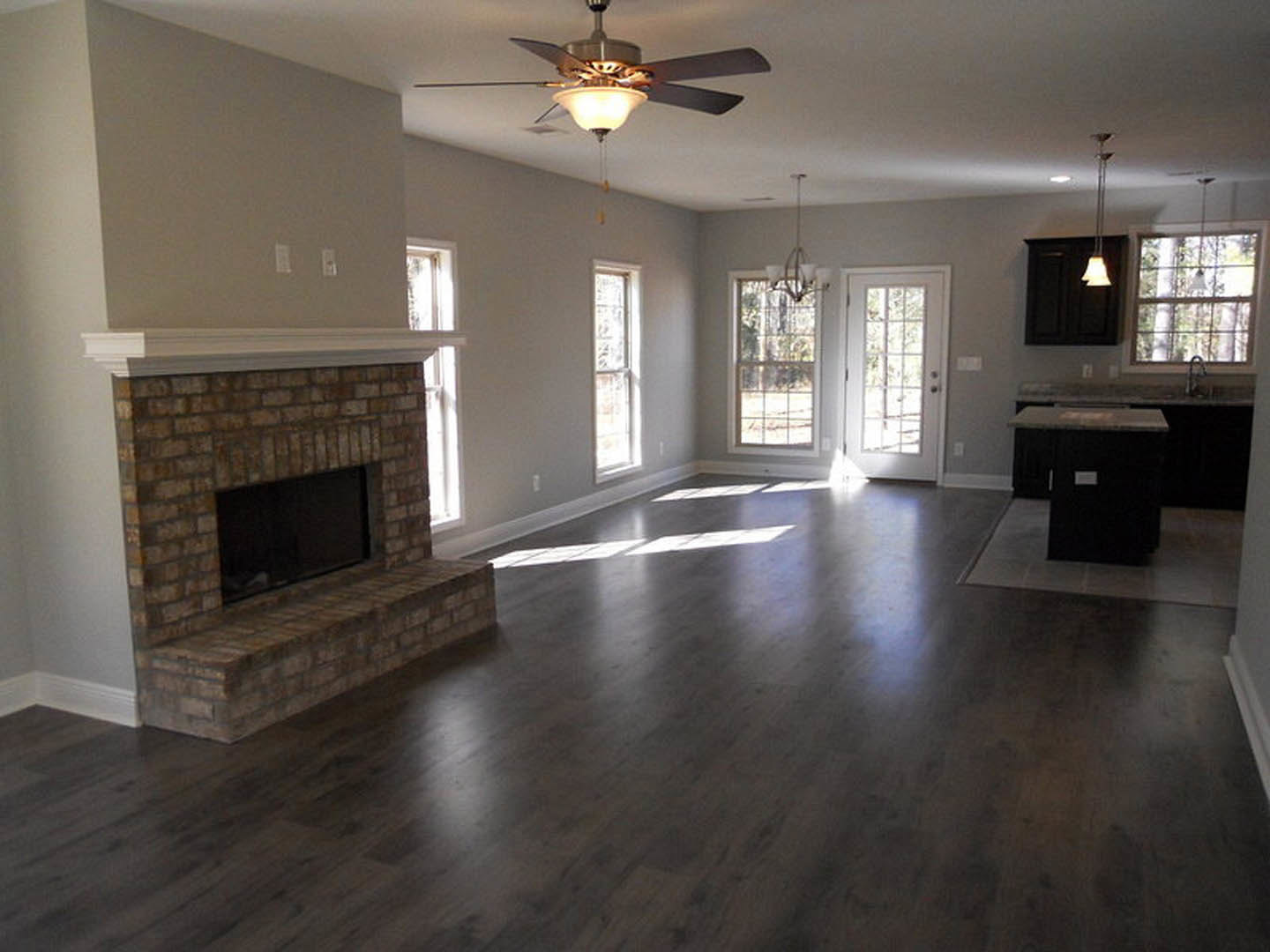 Living room featuring a brick hearth fireplace, ceiling fan with light, glass-paneled white door, and windows illuminating hardwood floors.