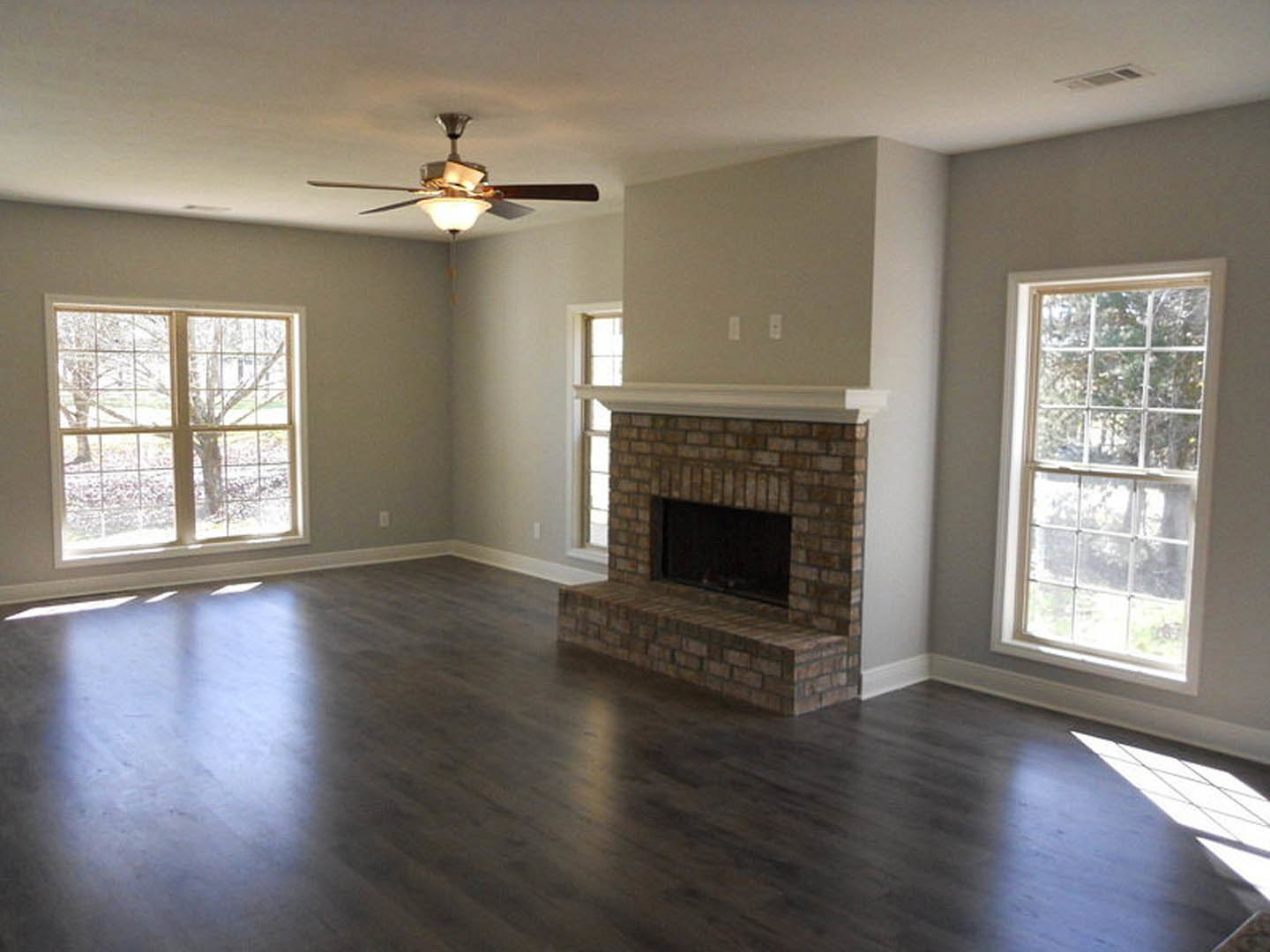Living room with wood flooring, white-framed fireplace, ceiling fan with light fixture, large window featuring multiple square panes and tree view