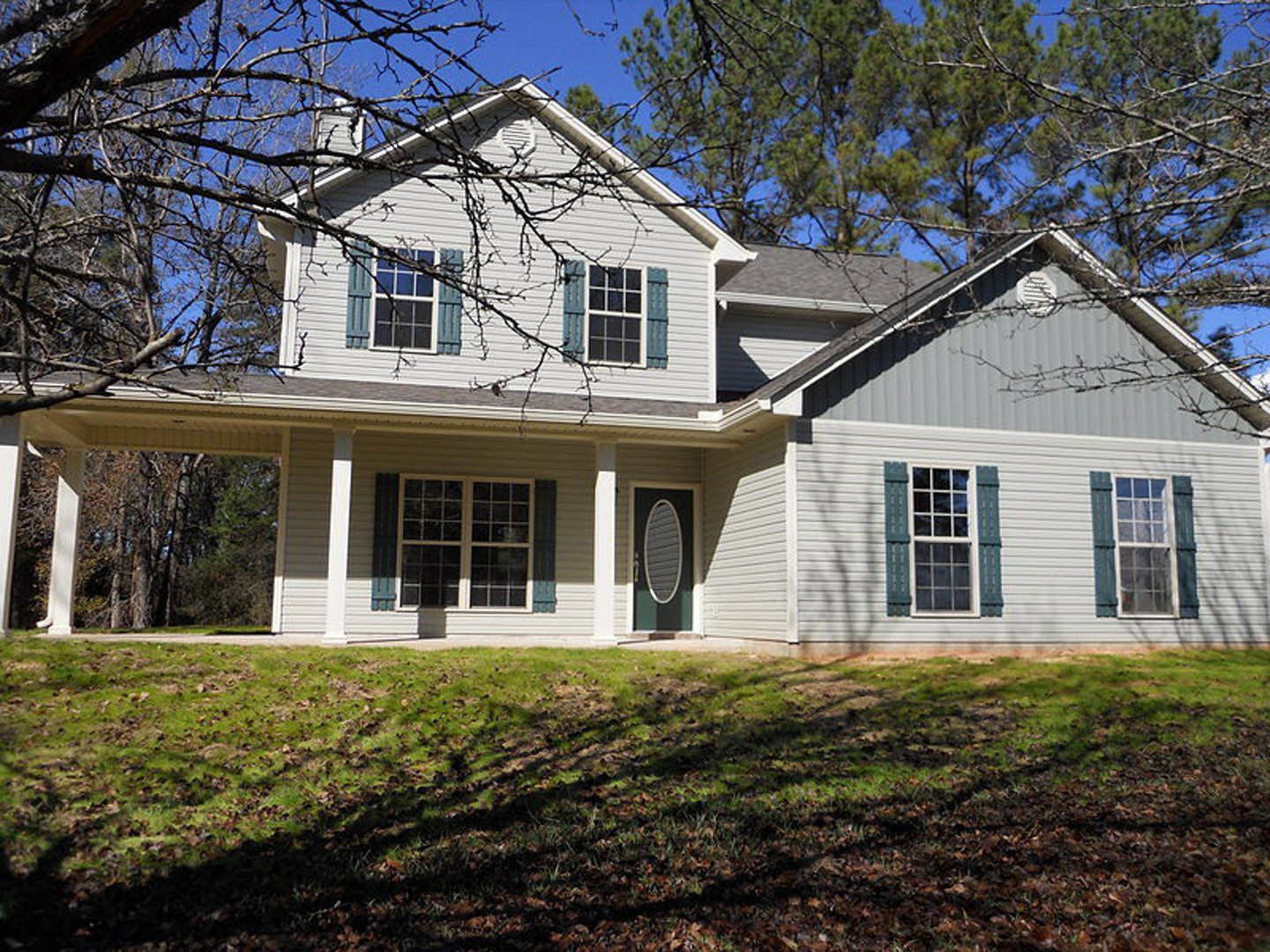 Two-story house with gray siding, green front door, white-trimmed windows, manicured lawn, and mature trees in the background