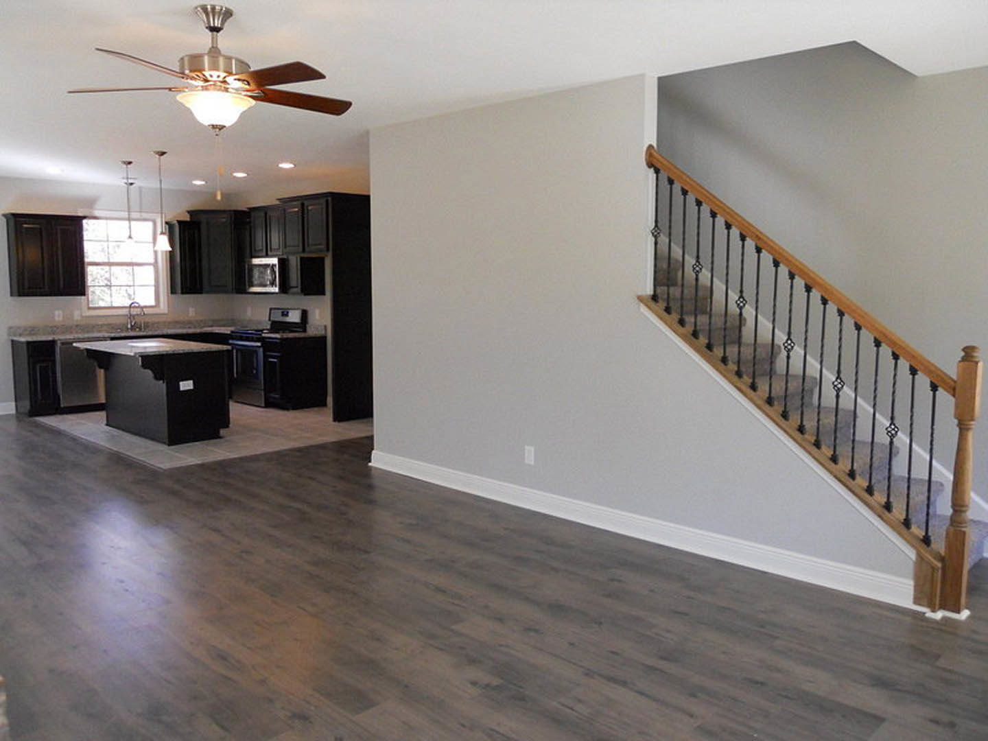 Open-concept kitchen and living room with wood flooring, marble-topped black island, metal-railed staircase, ceiling fan with light fixture, and large window.