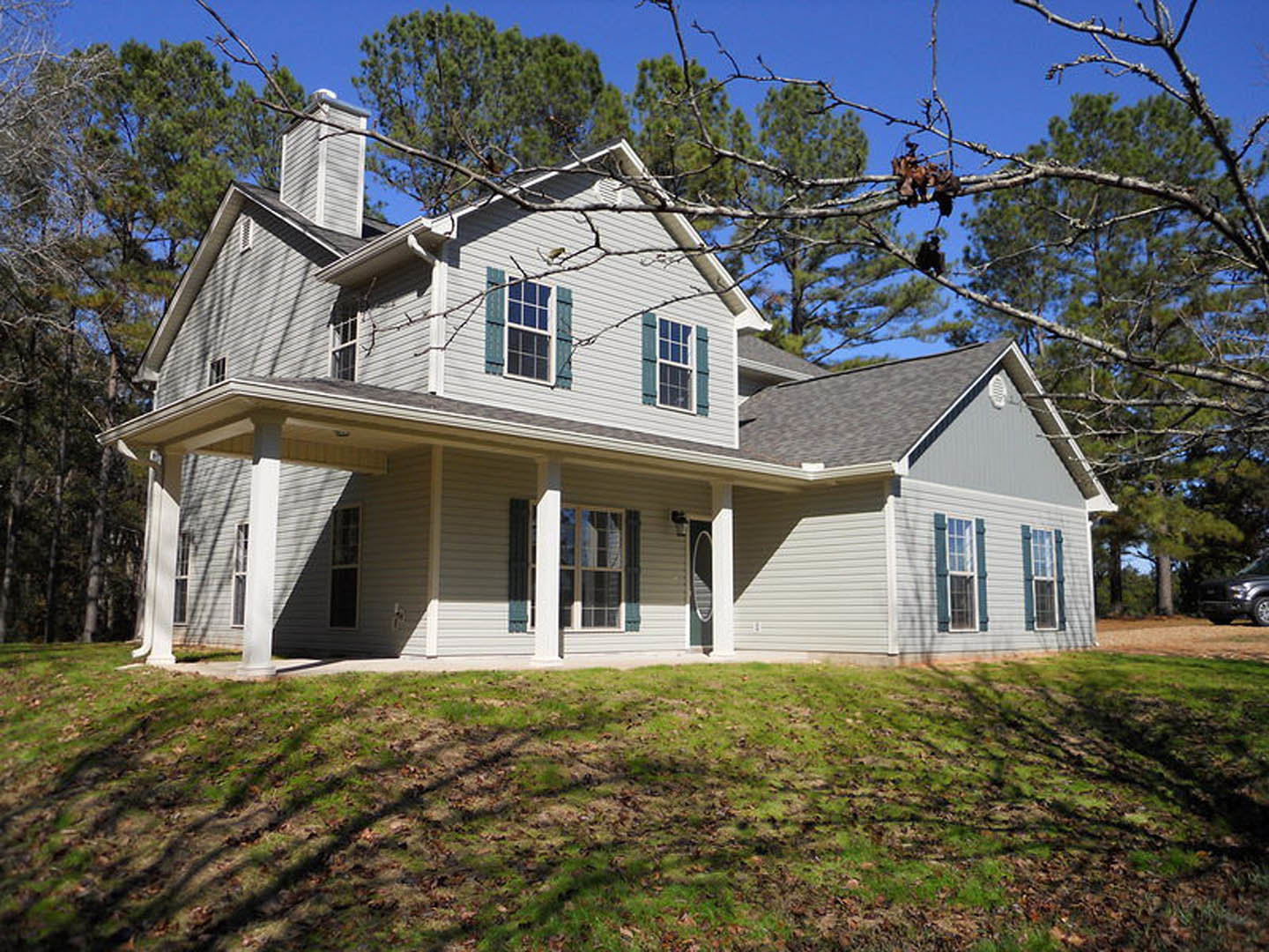 Two-story house with wide covered porch, white trim, large windows, tree branches overhead, green lawn scattered with fallen leaves