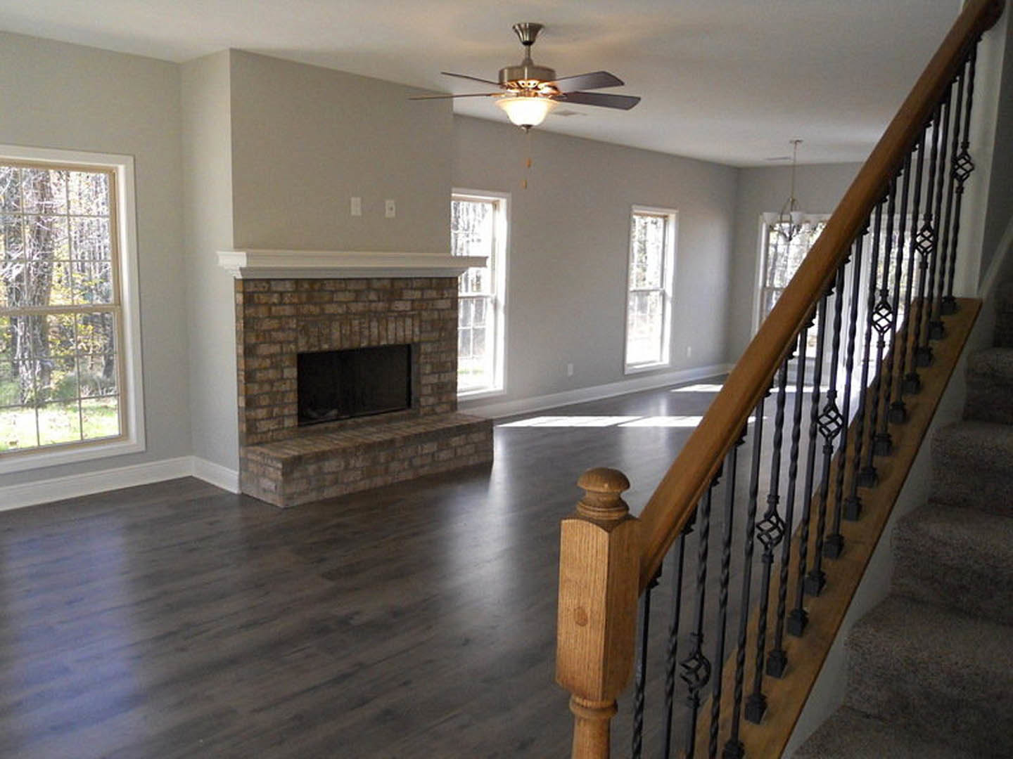 Living room with brick fireplace, wood staircase, large window showing trees, ceiling fan with light, hardwood floors, and crown molding; person walking in dimly lit space.