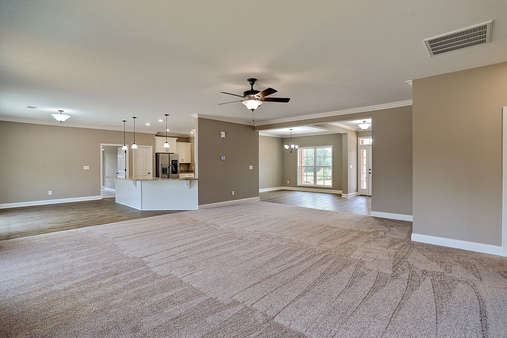 Spacious living room with neutral carpet flooring, ceiling fan and light fixture, large window overlooking outdoors, stainless steel refrigerator and faucet visible in adjacent