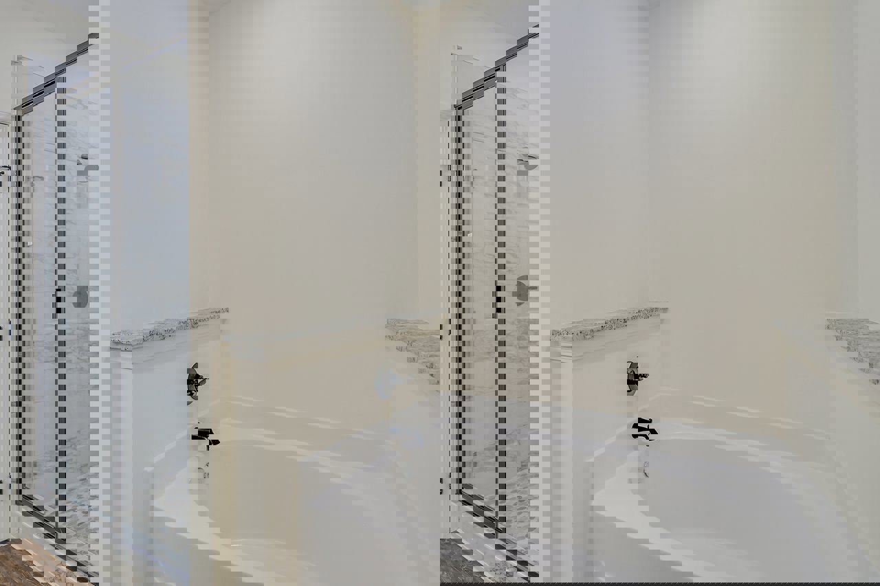 Freestanding white bathtub beside a glass shower door with marbled tile, black wall knob, and white countertop with black specks