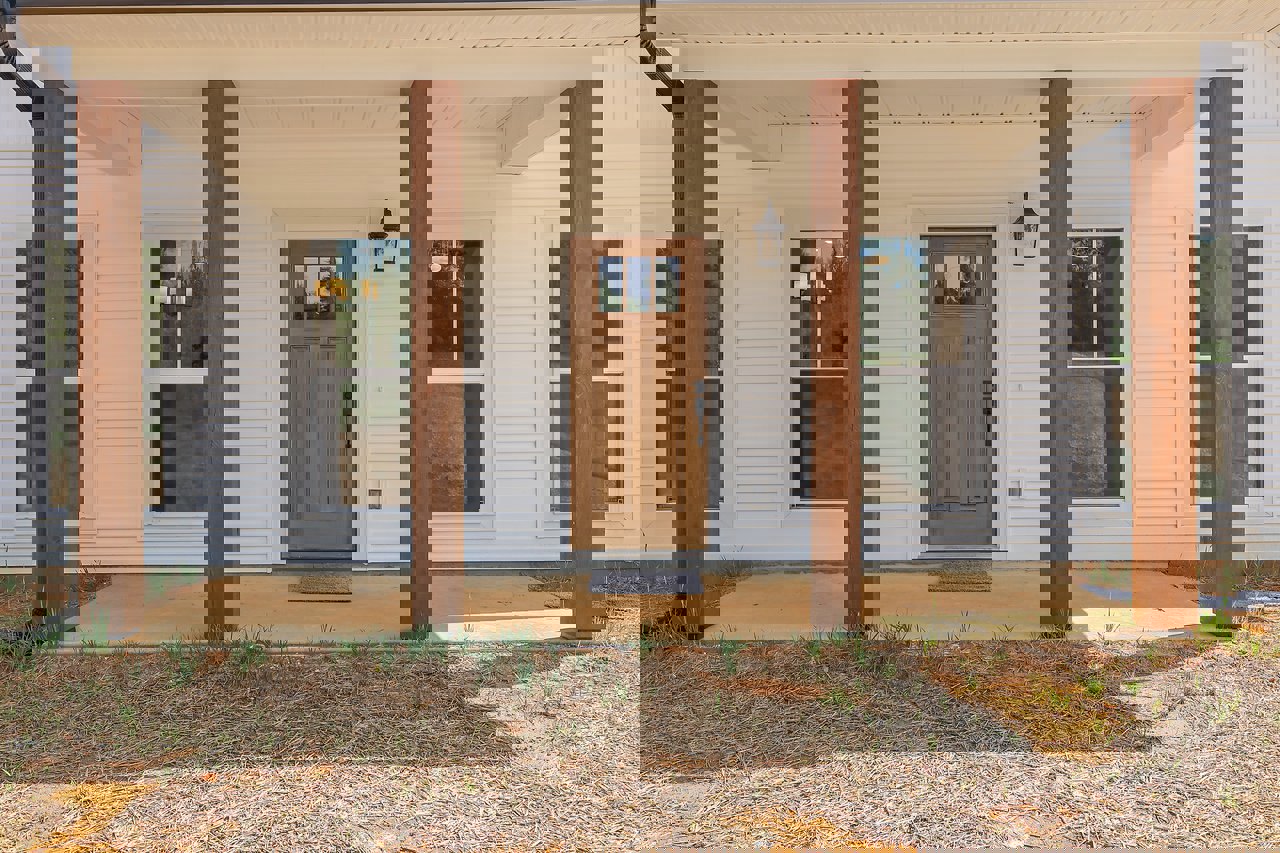 Front porch with brown door featuring glass window, white-framed window, wooden post against white tile wall, grey mat on concrete, grass and straw covering ground