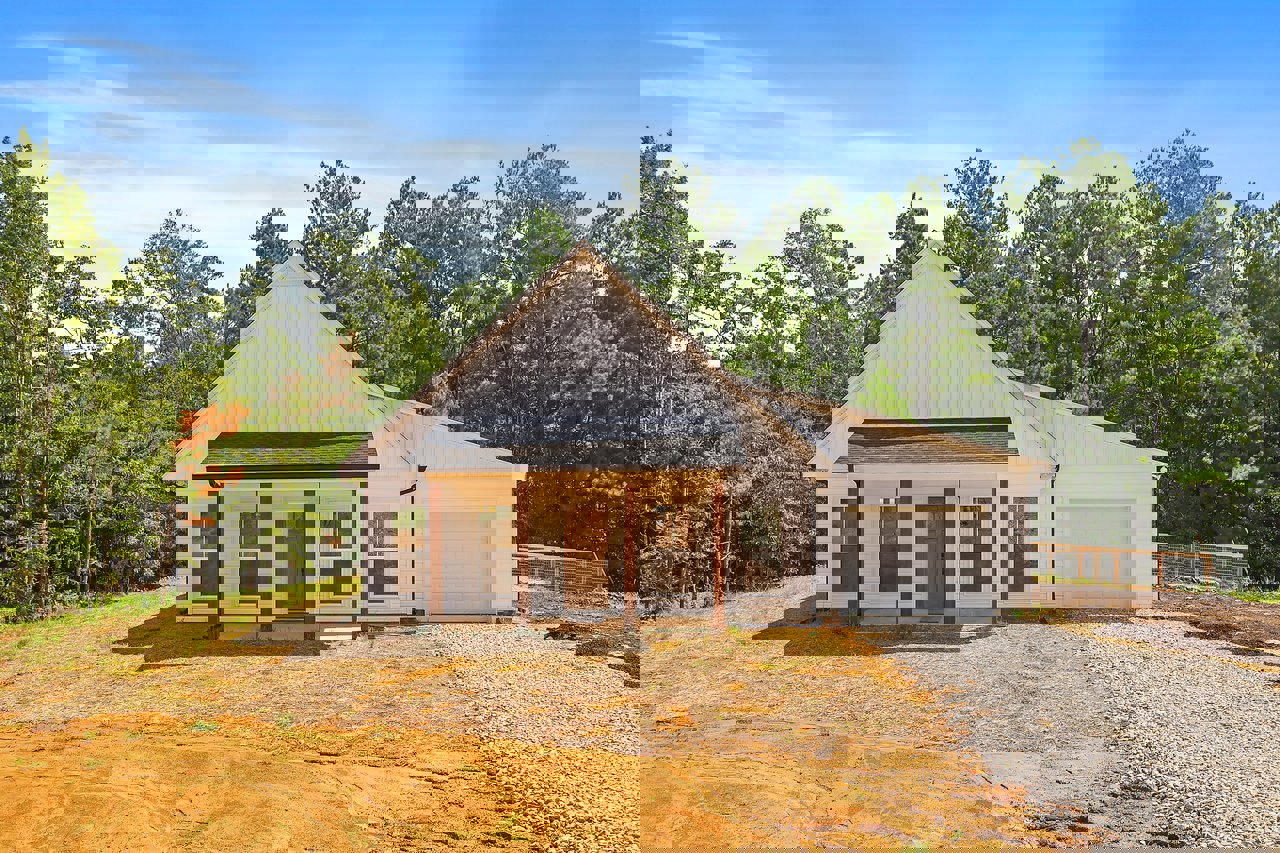 White cottage with gray gravel driveway, wire fence, covered porch, and trees under a partly cloudy blue sky; small shed visible in the background.