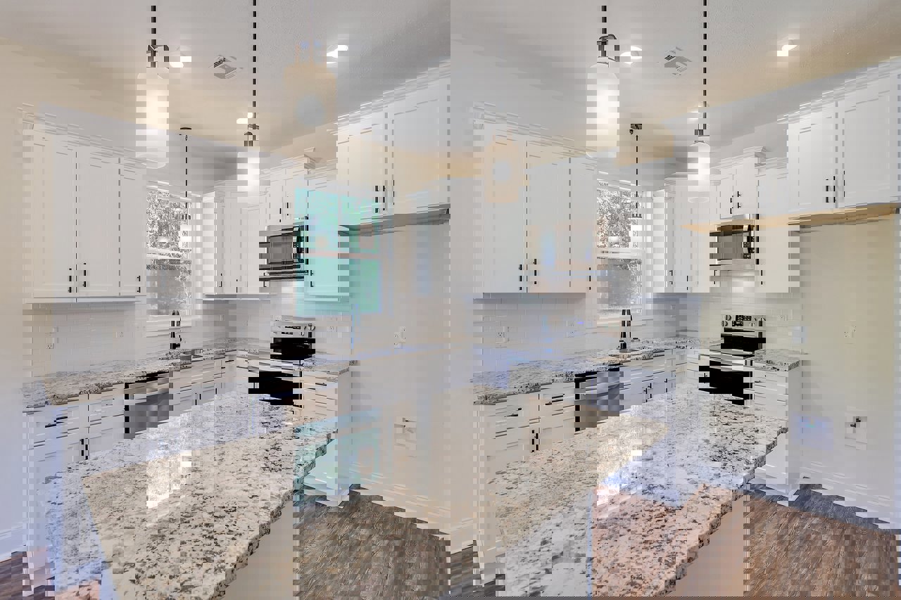 White kitchen cabinets, marble countertops with light reflection, stainless steel oven and stove, built-in microwave with glass doors, sink, and modern light fixture near window.