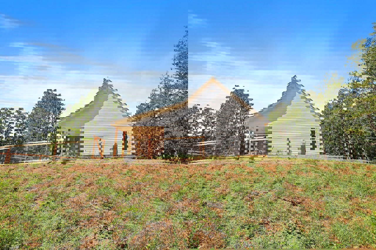 White farmhouse with gabled roof, wooden wire fence enclosing grassy yard, mature trees and blue sky in background