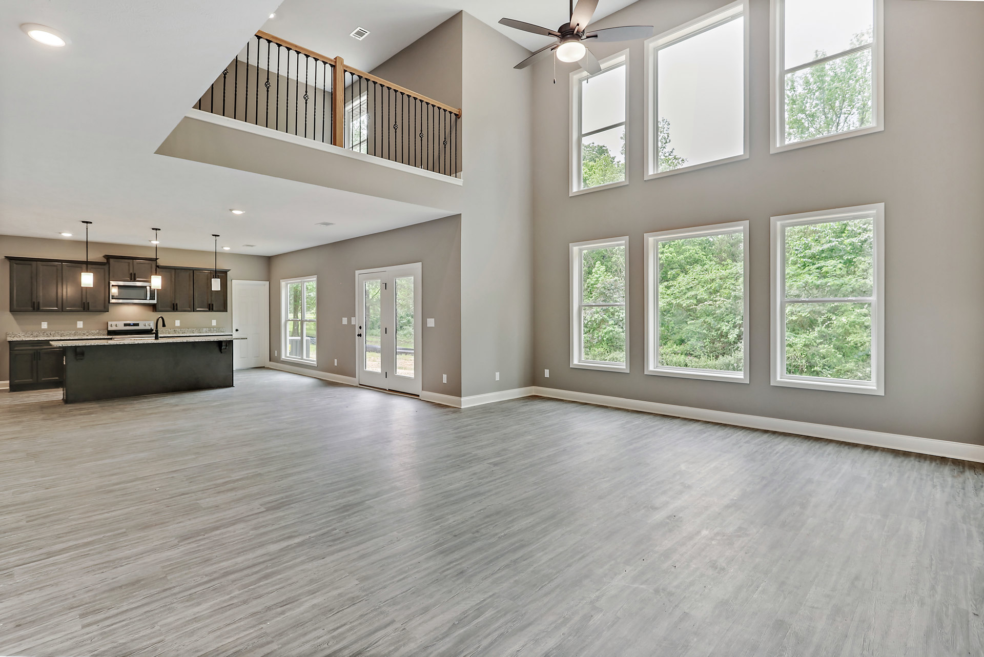 Open-concept room featuring wood flooring, modern kitchen with white cabinetry, large ceiling fan with light, expansive windows overlooking trees, and glass door leading outside