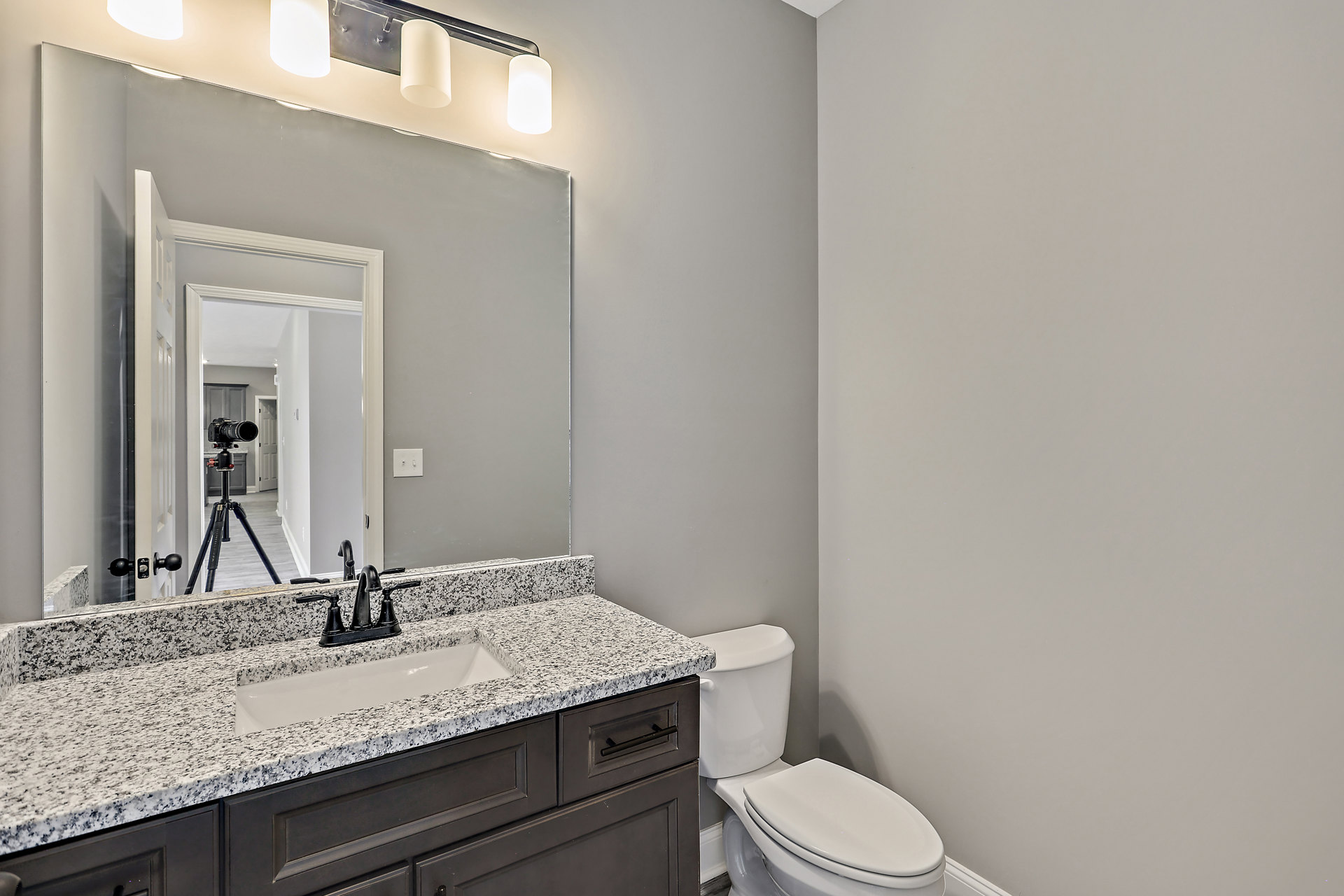 White toilet with closed lid beside a marbled countertop and black faucet, white cylindrical light fixture overhead, light tile flooring and neutral cabinetry visible.