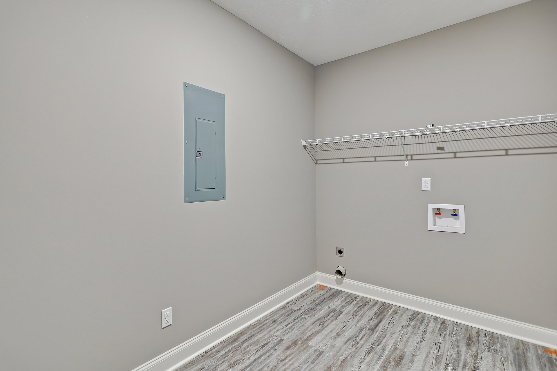 White plaster wall with mounted shelf, wood floor featuring exposed drain pipe, grey metal utility box with square door, white box with red and blue valves, camera placed on metal