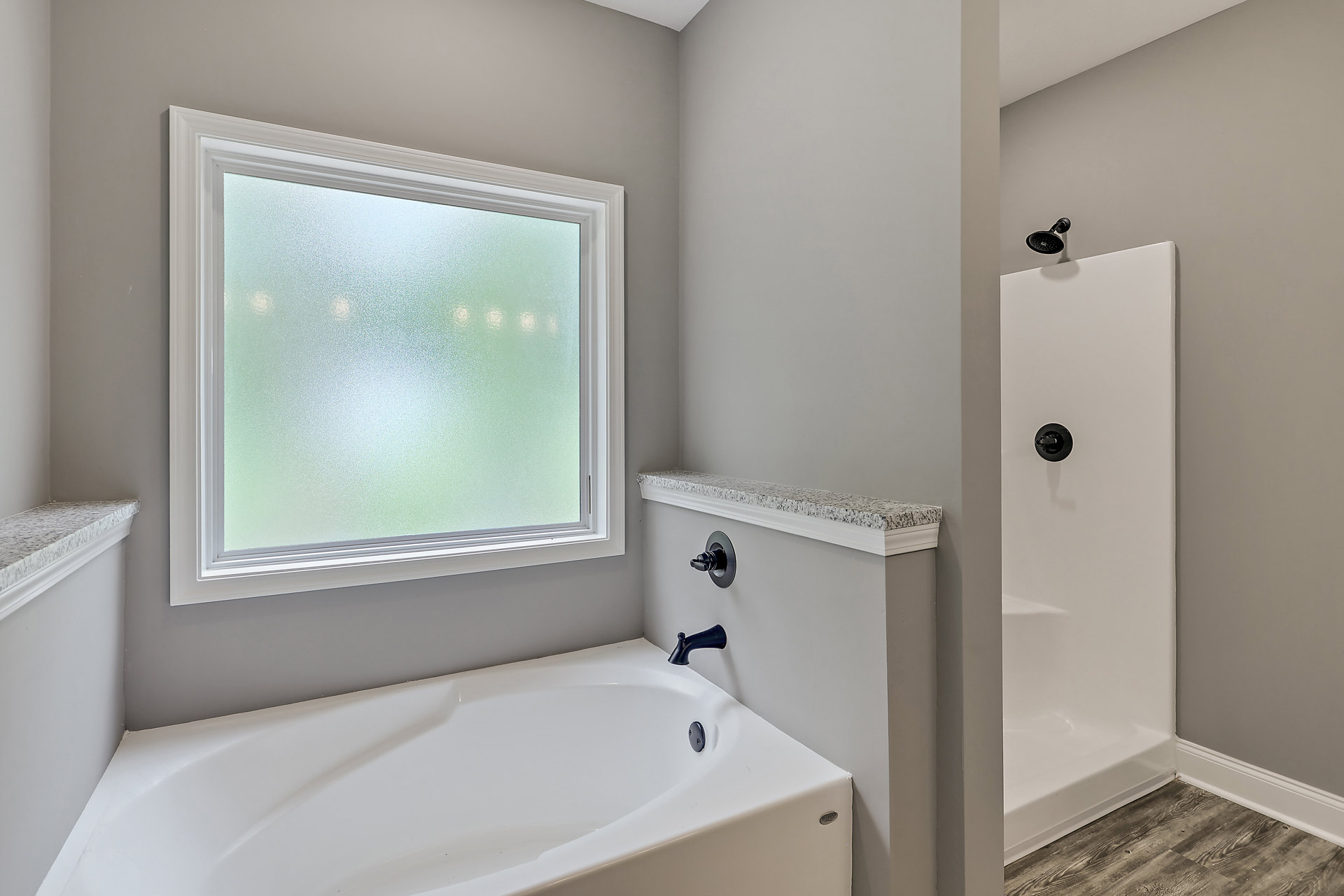 White freestanding bathtub with black faucet beside frosted glass window, wall-mounted shower head, light gray tile walls, and minimal modern bathroom fixtures