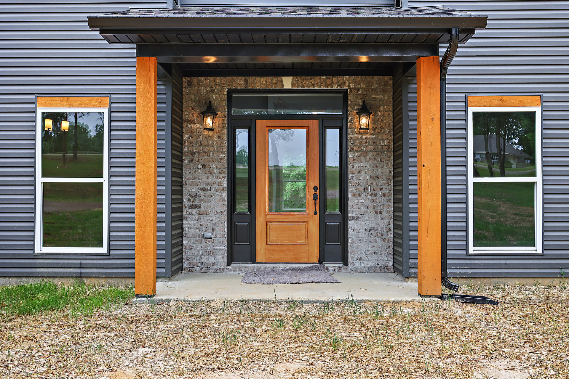 Wide front porch with brick walls, dark wood front door, large windows overlooking green lawn and trees, black metal light fixture, grey carpeted entry area