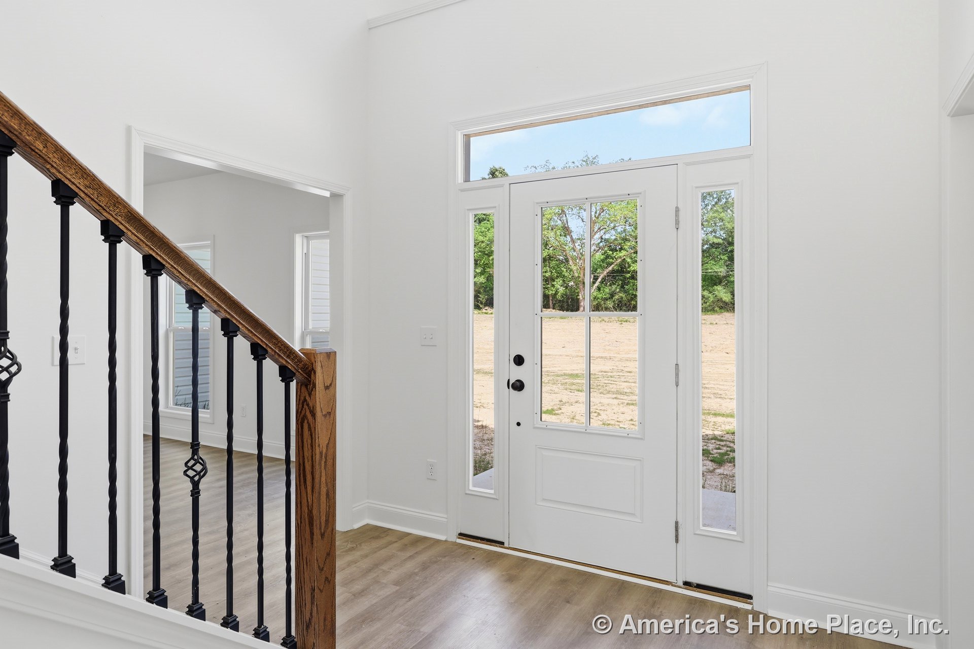 White front door with glass sidelights and transom window, adjacent to staircase featuring wood handrail and black iron balusters; light wood flooring and white interior trim