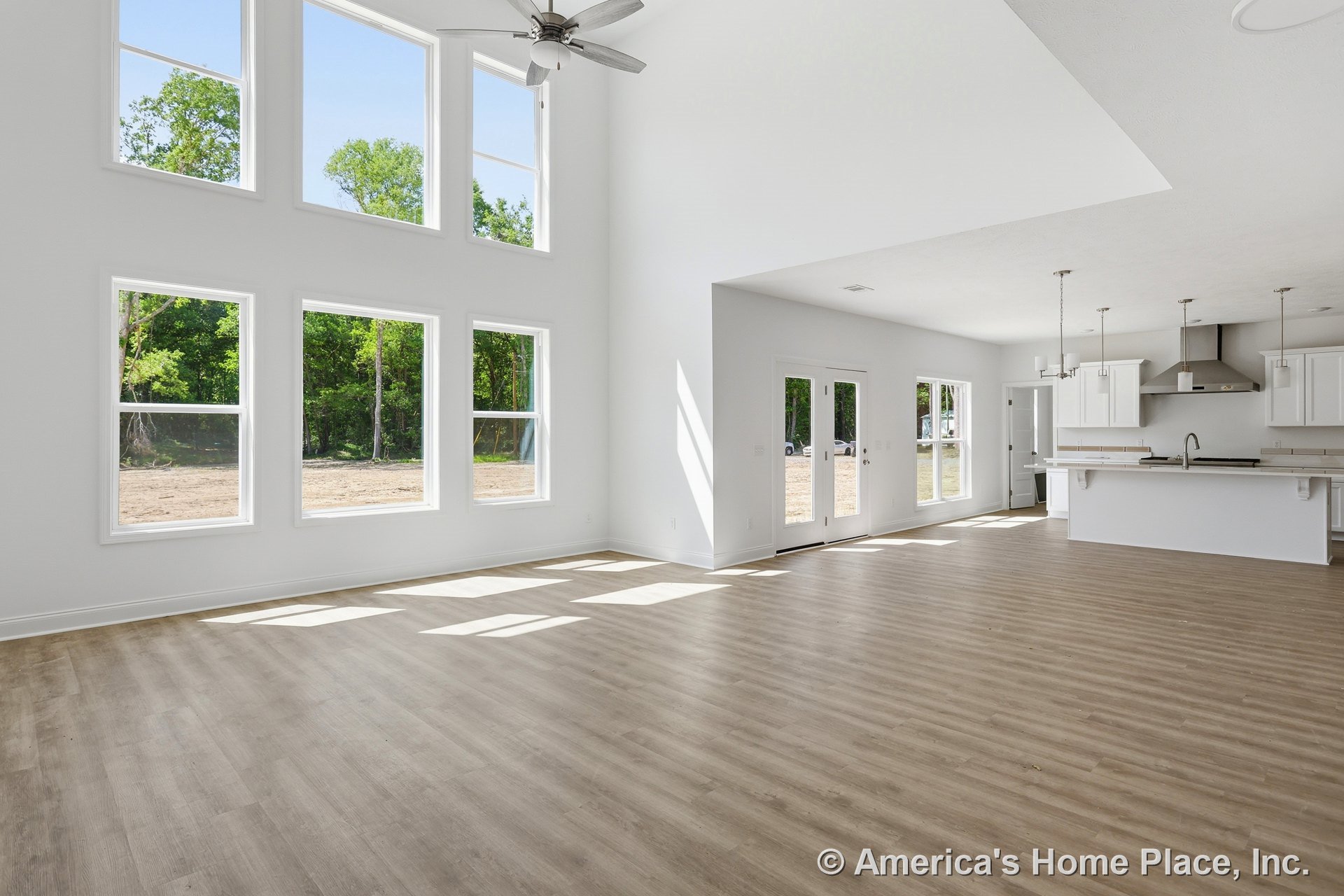 Double-height ceiling living area with expansive windows, open floor plan connecting to modern kitchen featuring white cabinetry, pendant lights above island countertop, light wood