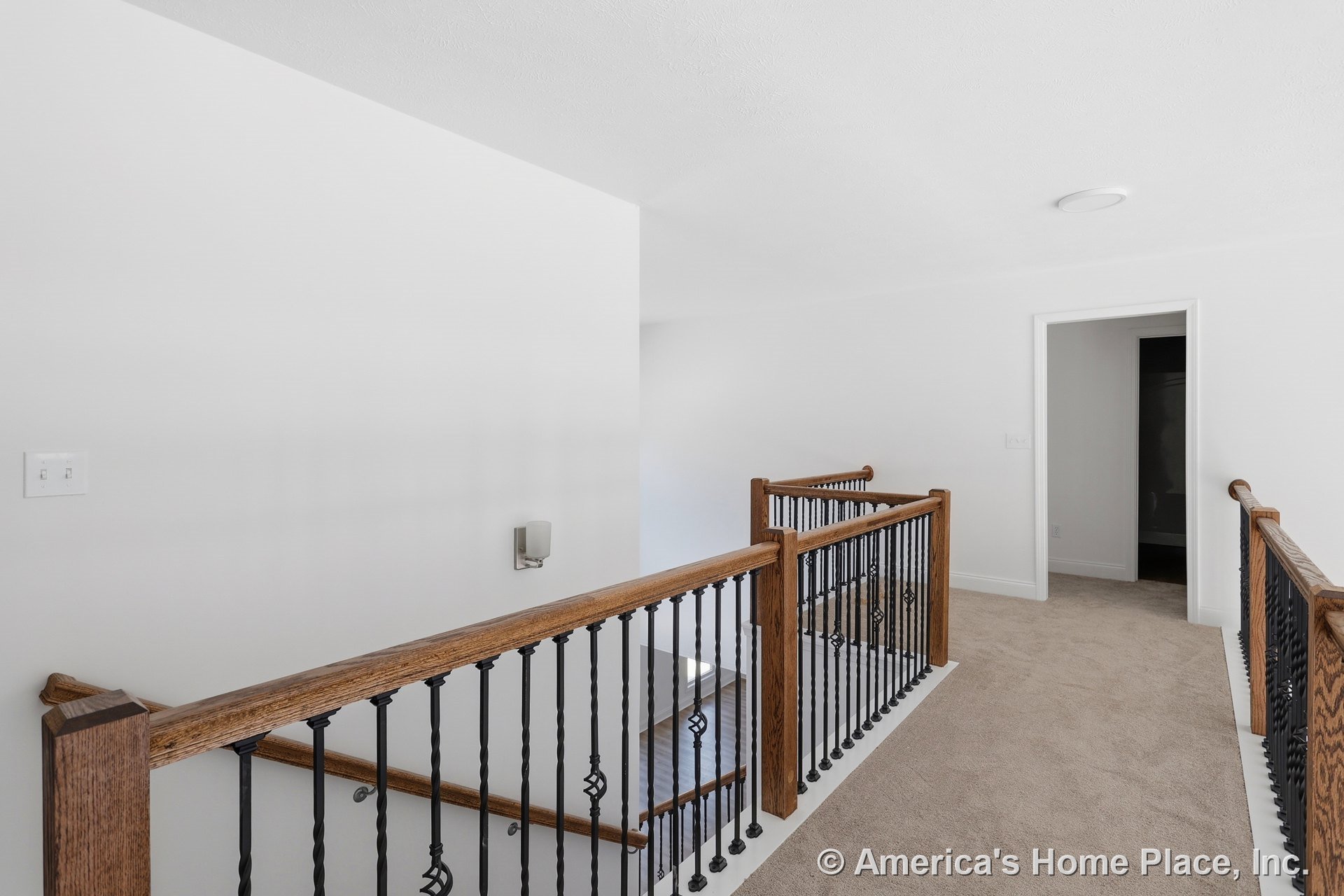 Upstairs hallway with carpet flooring, wooden stair railing accented by decorative black iron balusters, white walls, ceiling light fixture, wall sconce, and trimmed doorway.