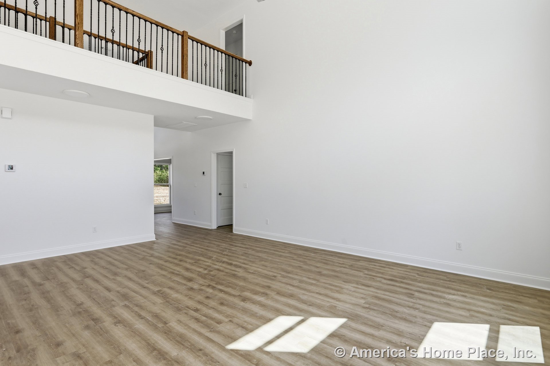 Spacious open living room with light wood plank flooring, white walls and trim, high ceiling, white-painted balcony featuring iron balusters, multiple interior doors, and abundant