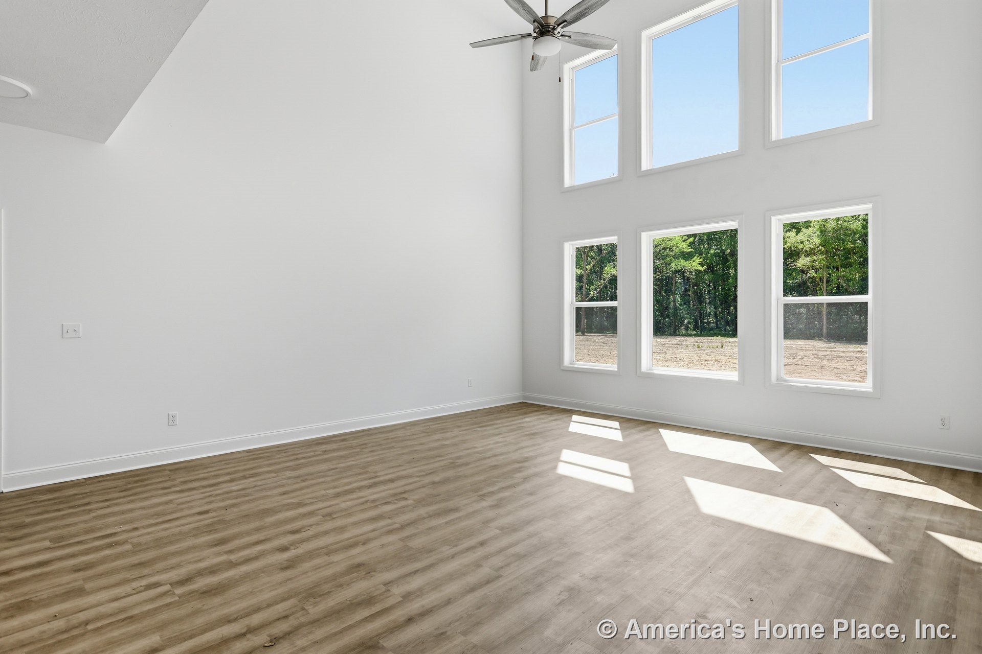 Double-height living room with tall ceiling, ceiling fan, five expansive windows, white walls, light wood plank flooring, and baseboard trim flooded with natural light.