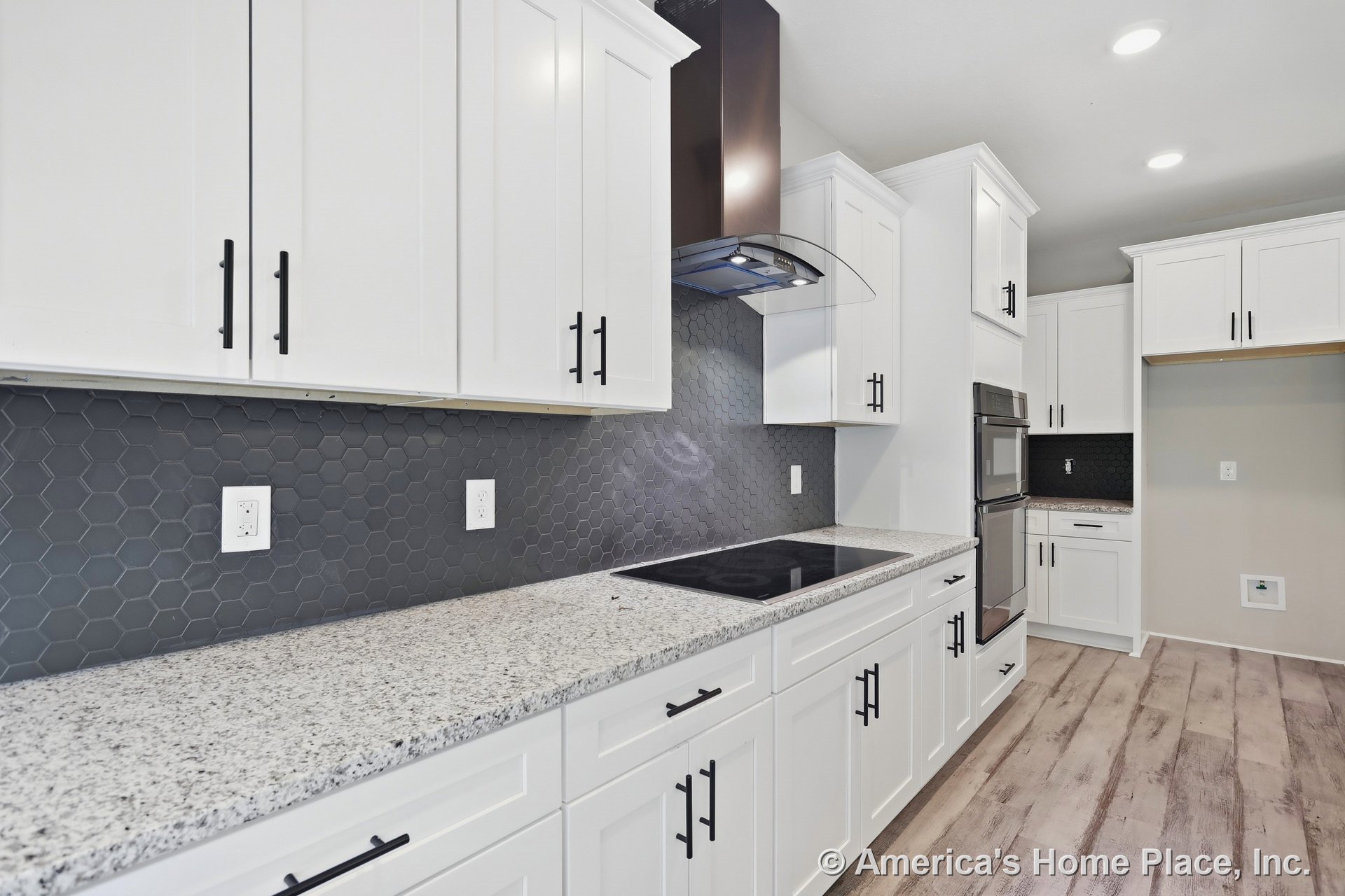 White shaker cabinets with brushed hardware, granite countertops, dark hexagonal tile backsplash, electric cooktop beneath a modern vent hood, double wall oven, built-in