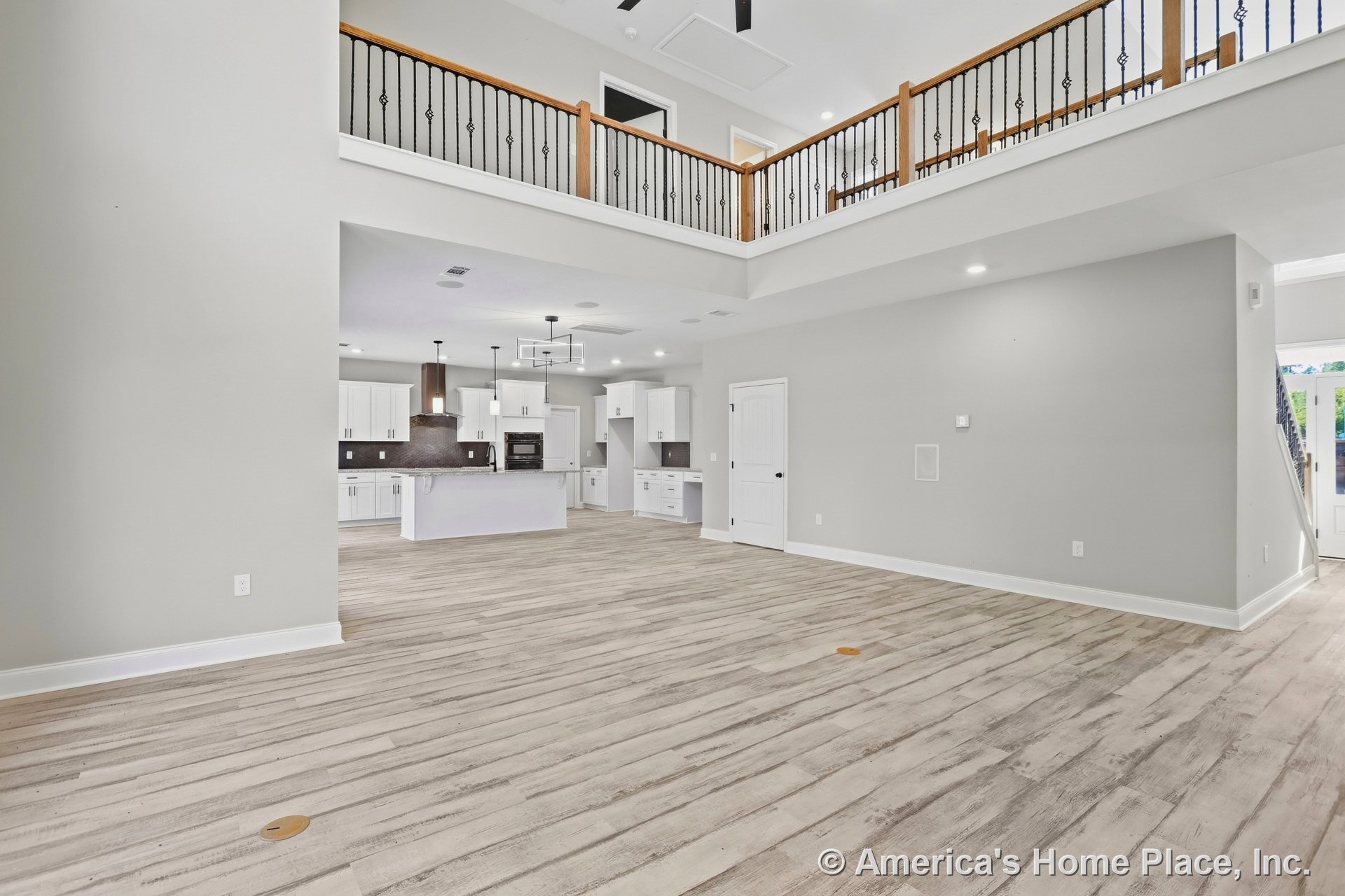 Open-concept living area with light wood-look plank flooring, modern kitchen featuring white cabinetry and dark tile backsplash, iron and wood stair railing, recessed ceiling
