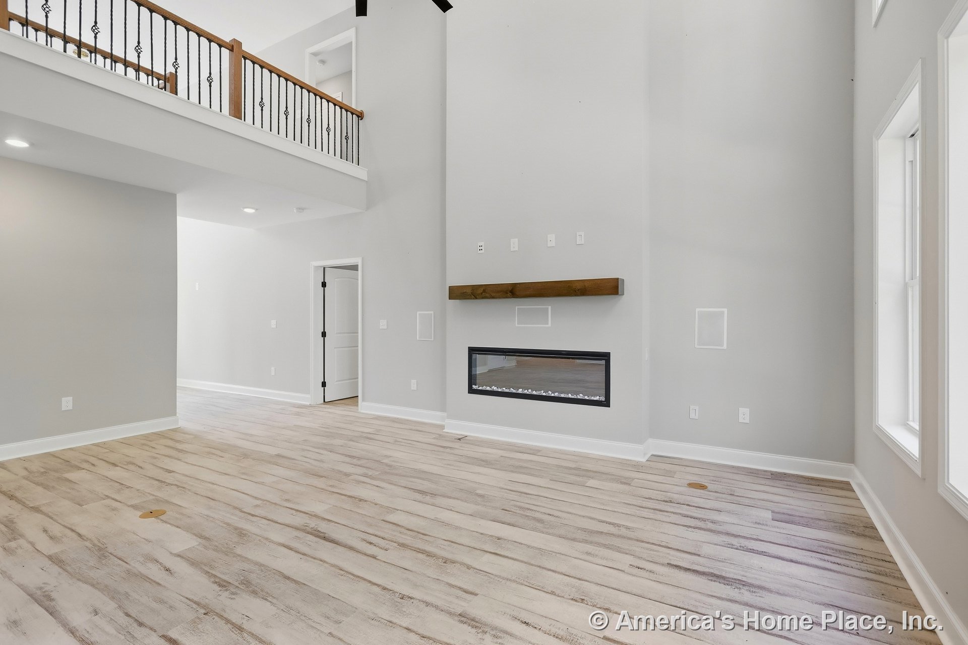 Double-height living room with expansive window wall, modern linear fireplace framed by a wood mantel, light-toned plank flooring, iron railing balcony overlooking space, white