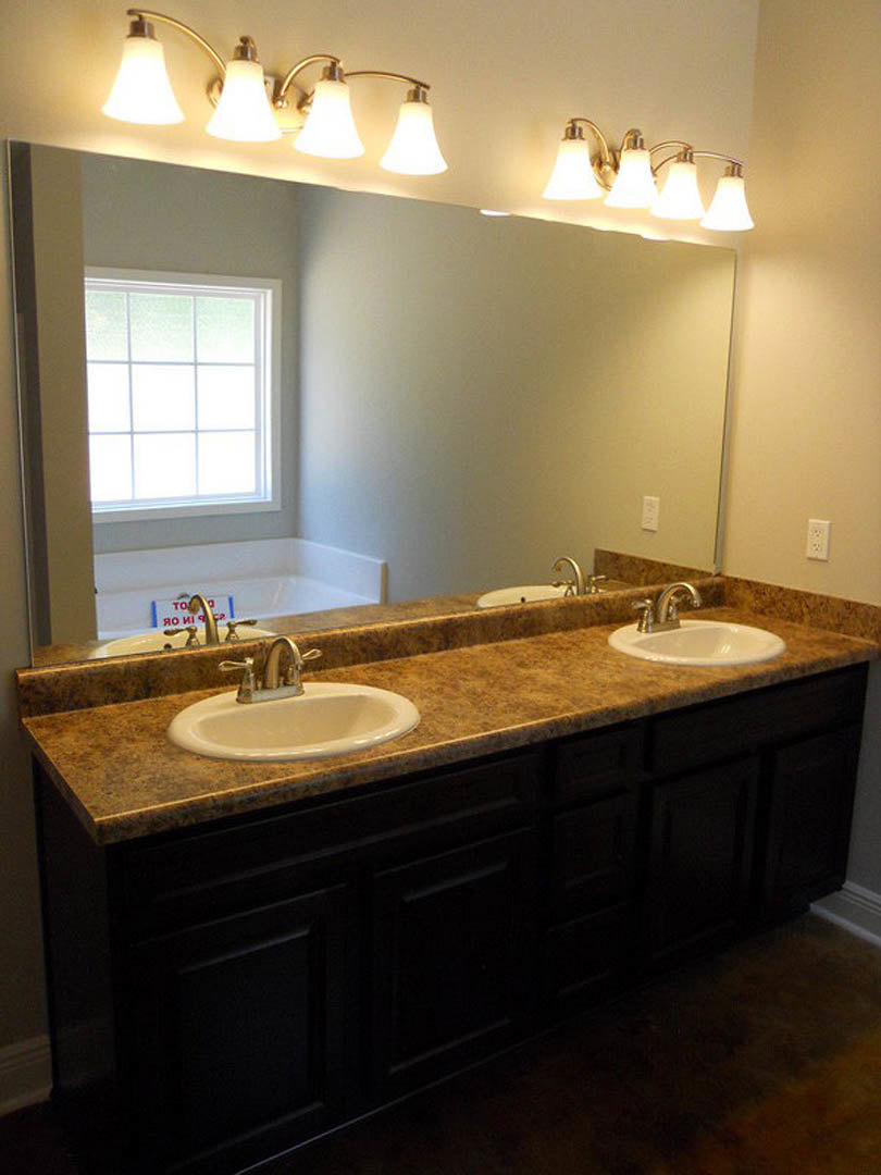 Bathroom with two undermount sinks set in a stone countertop, large framed mirror above, three-light fixture mounted on wall, chrome faucets, white tile backsplash, window with