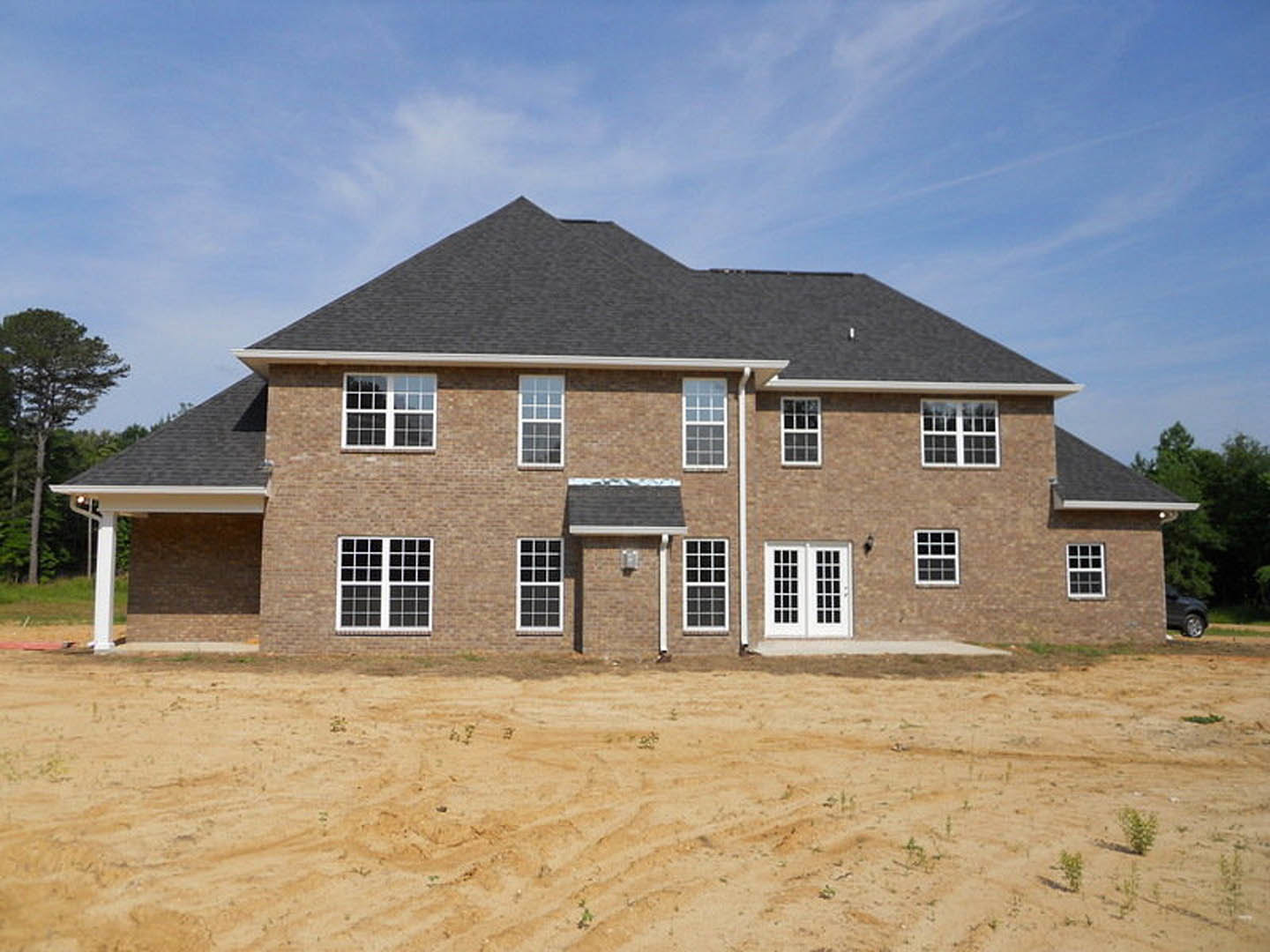 Brick house with black roof under construction, surrounded by dirt field, multiple square windows visible, trees in background, cloudy sky overhead