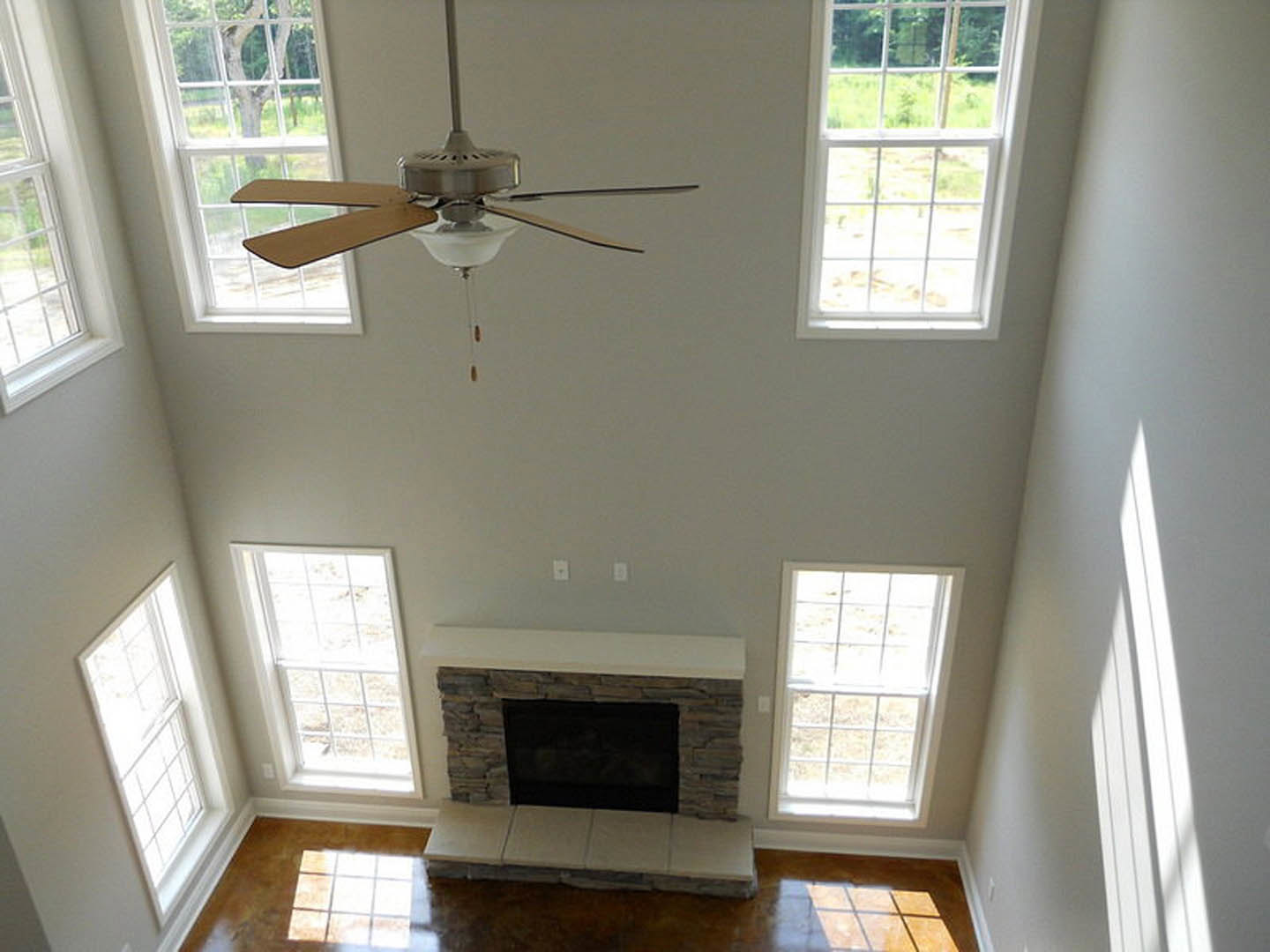 Ceiling fan mounted on white ceiling above living room with large windows, white-framed fireplace with built-in shelf, light wood flooring, and white tile accents