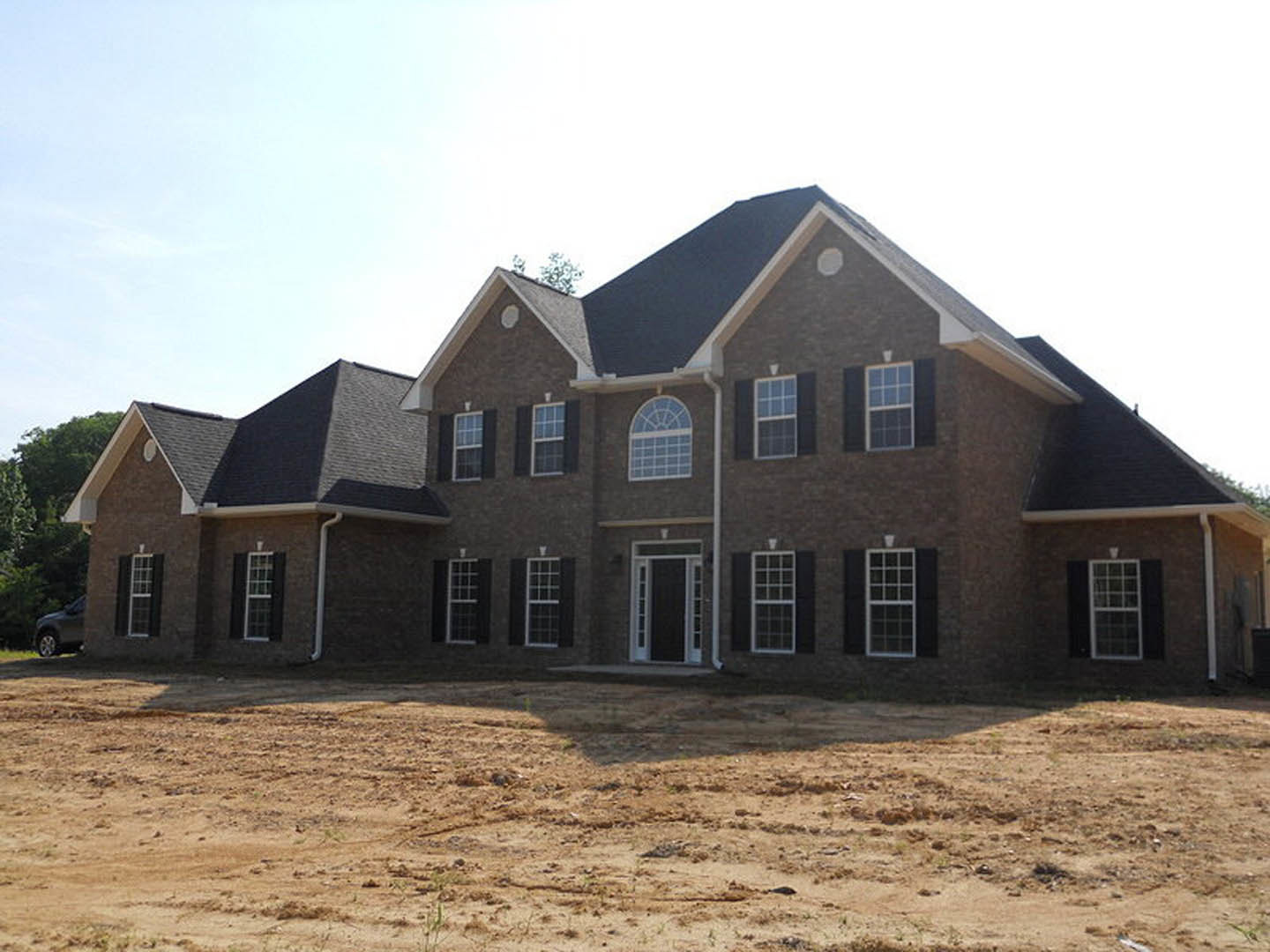 Partially built house with white door and white-trimmed grid windows, surrounded by dirt field and sparse trees under a clear sky