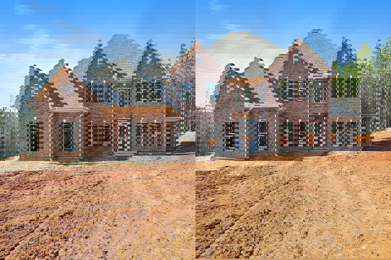 Partially built brick house with brown door and glass panes, surrounded by dirt field and tire tracks, trees in background under cloudy sky