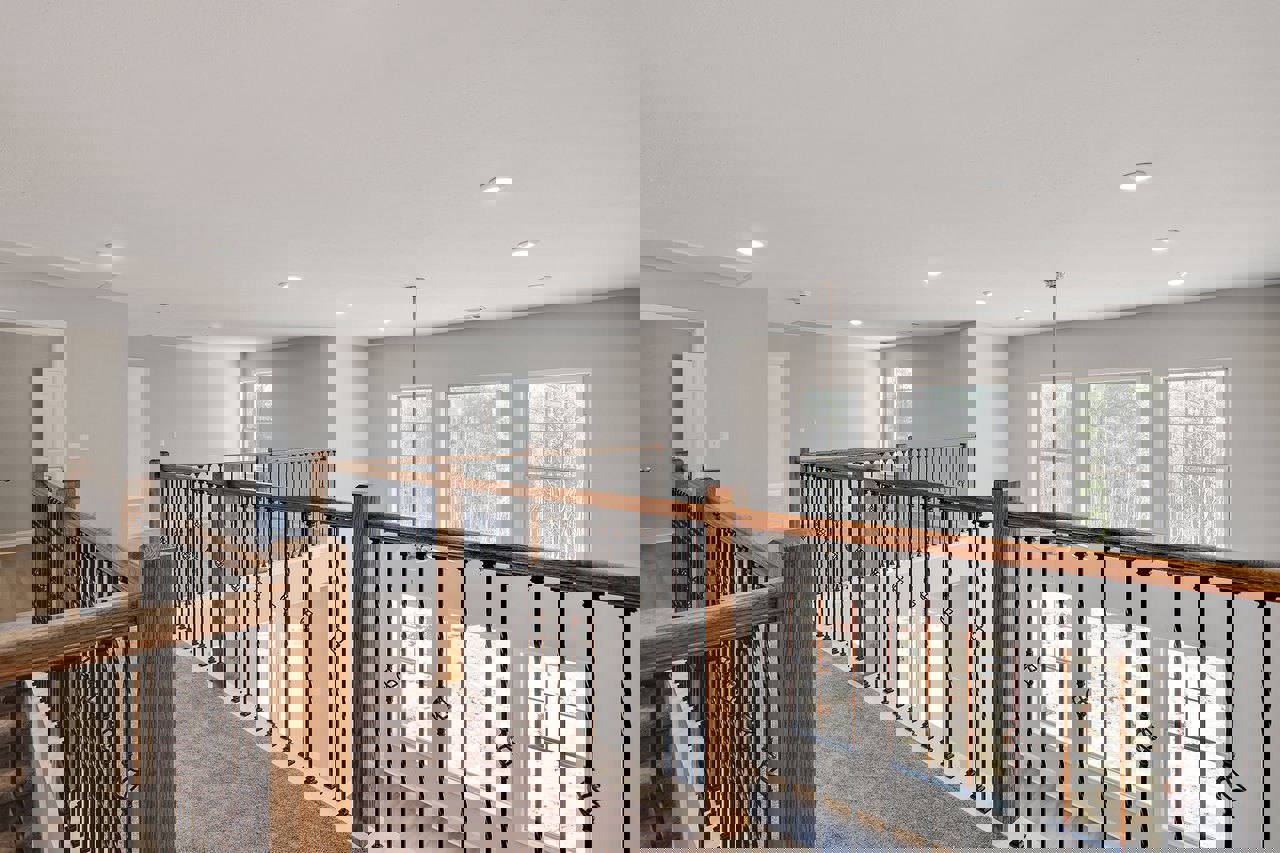 Wood staircase with dark railing, white plaster walls, recessed ceiling lights, large window overlooking green trees.