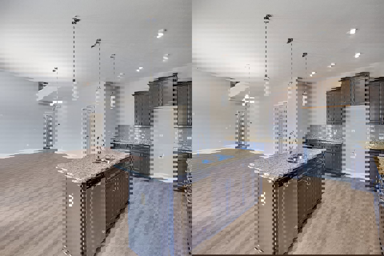 Spacious kitchen featuring a large central island with built-in sink and dishwasher, wood flooring, silver refrigerator with sticker, white walls accented by a black pole, white