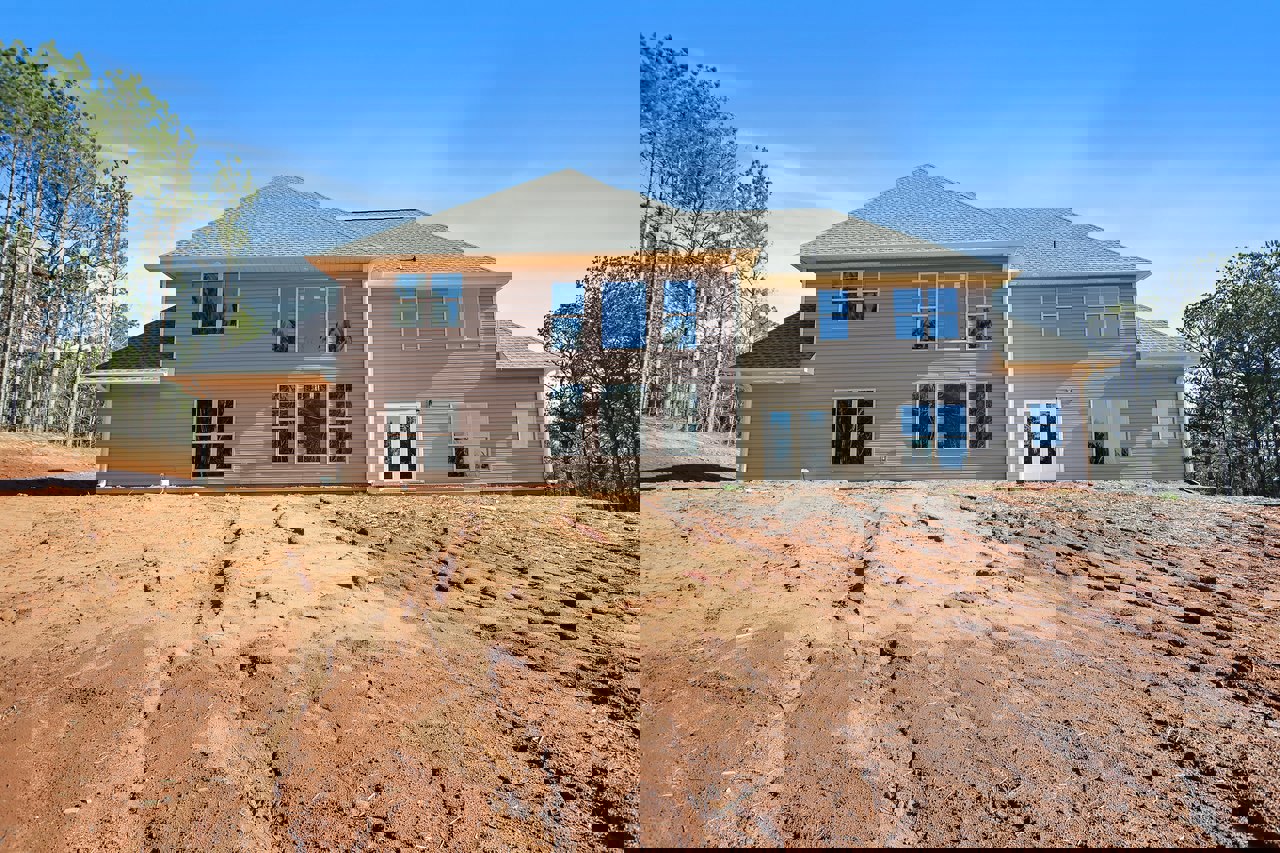 Modern home with multiple large windows, blue front door featuring a sticker, dirt patch and hill in the foreground, surrounded by mature trees under a clear blue sky