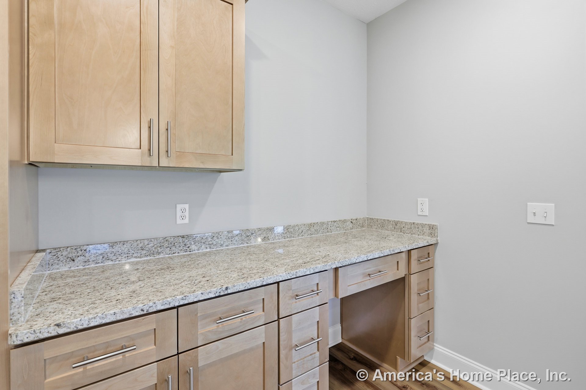 Built-in workspace with light wood upper cabinets, granite countertop and matching backsplash, multiple drawers, desk cutout, light gray walls, and wood-look flooring.