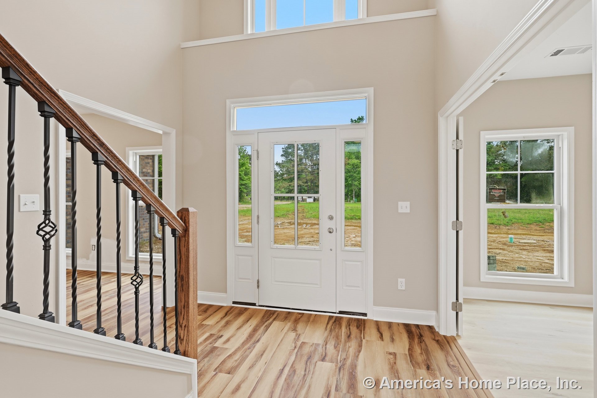 Double-height foyer with white trim and molding, glass panel front door, tall windows, light wood-look flooring, and staircase featuring wood railing and iron balusters.