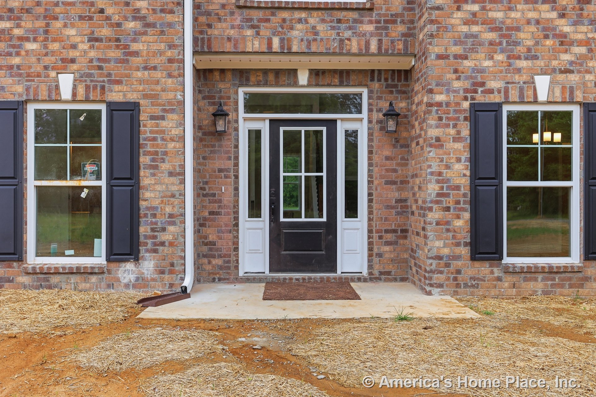 Brick facade with black front door featuring glass panels, white trim and sidelights, lantern-style wall lights on either side, double windows with black shutters, and concrete