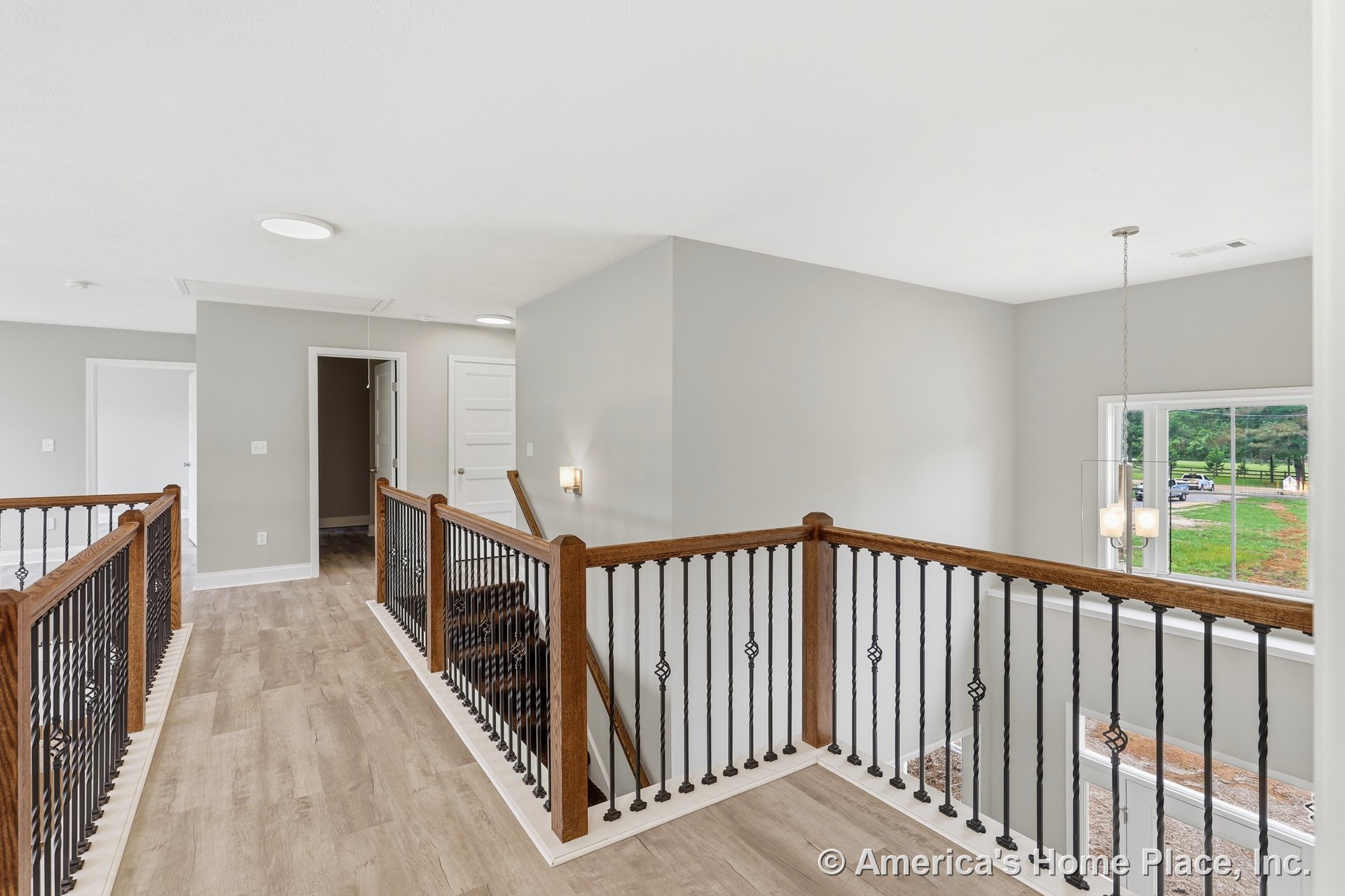 Upstairs landing with wood and iron stair railings, light wood plank flooring, large window framed in clear trim, modern pendant light fixture, neutral gray walls, and white