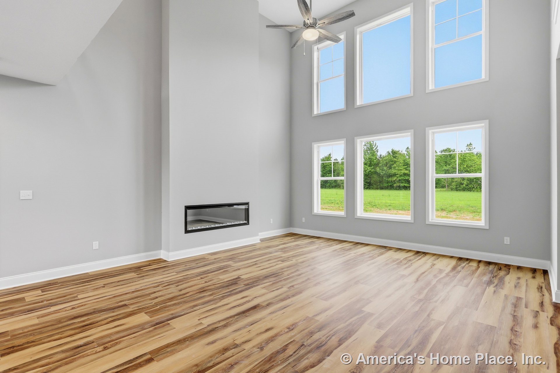 Double-height living room with expansive stacked windows, modern linear fireplace set against gray painted walls, wood-look plank flooring, ceiling fan with light, and crisp white