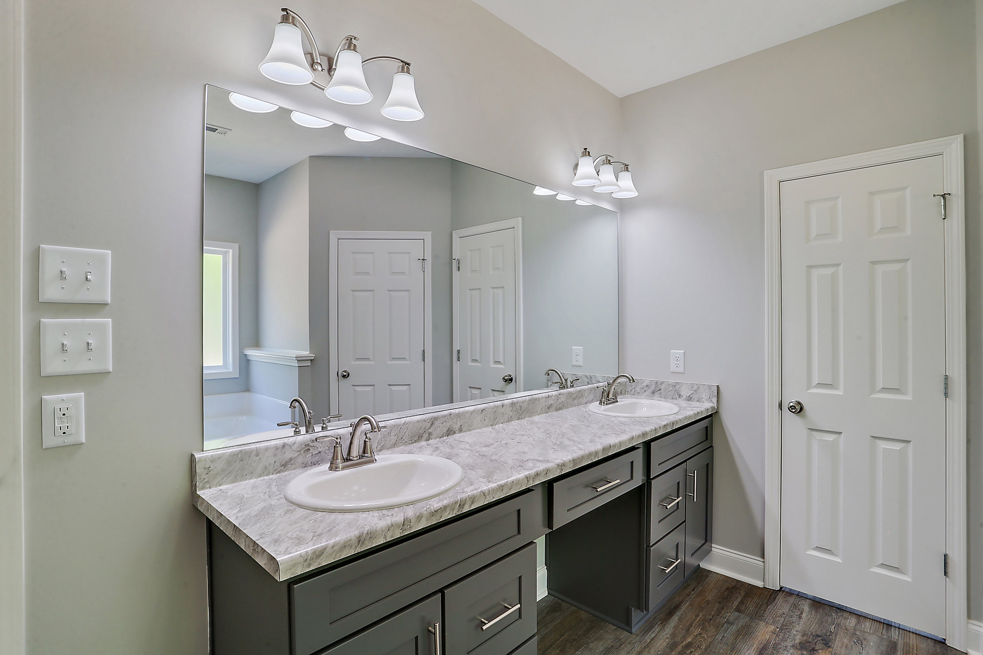 Bathroom with expansive mirror above white countertop, chrome faucet, white cabinetry, and silver door knob on adjacent white door; visible wall outlet and light switch.