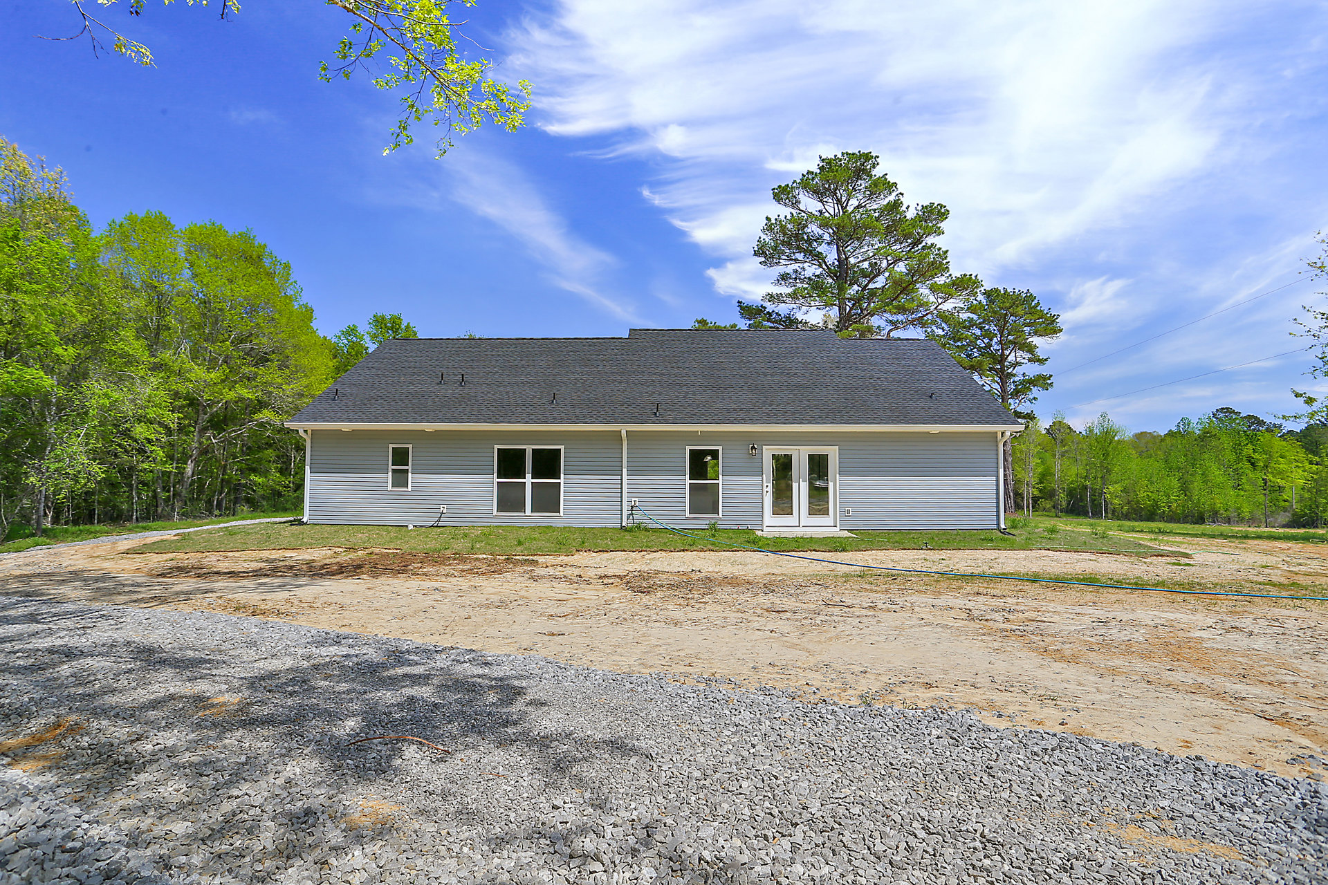 Gravel driveway bordered by green lawn and mature trees leading to a cottage-style house with white double doors, glass windows, and a gabled roof.