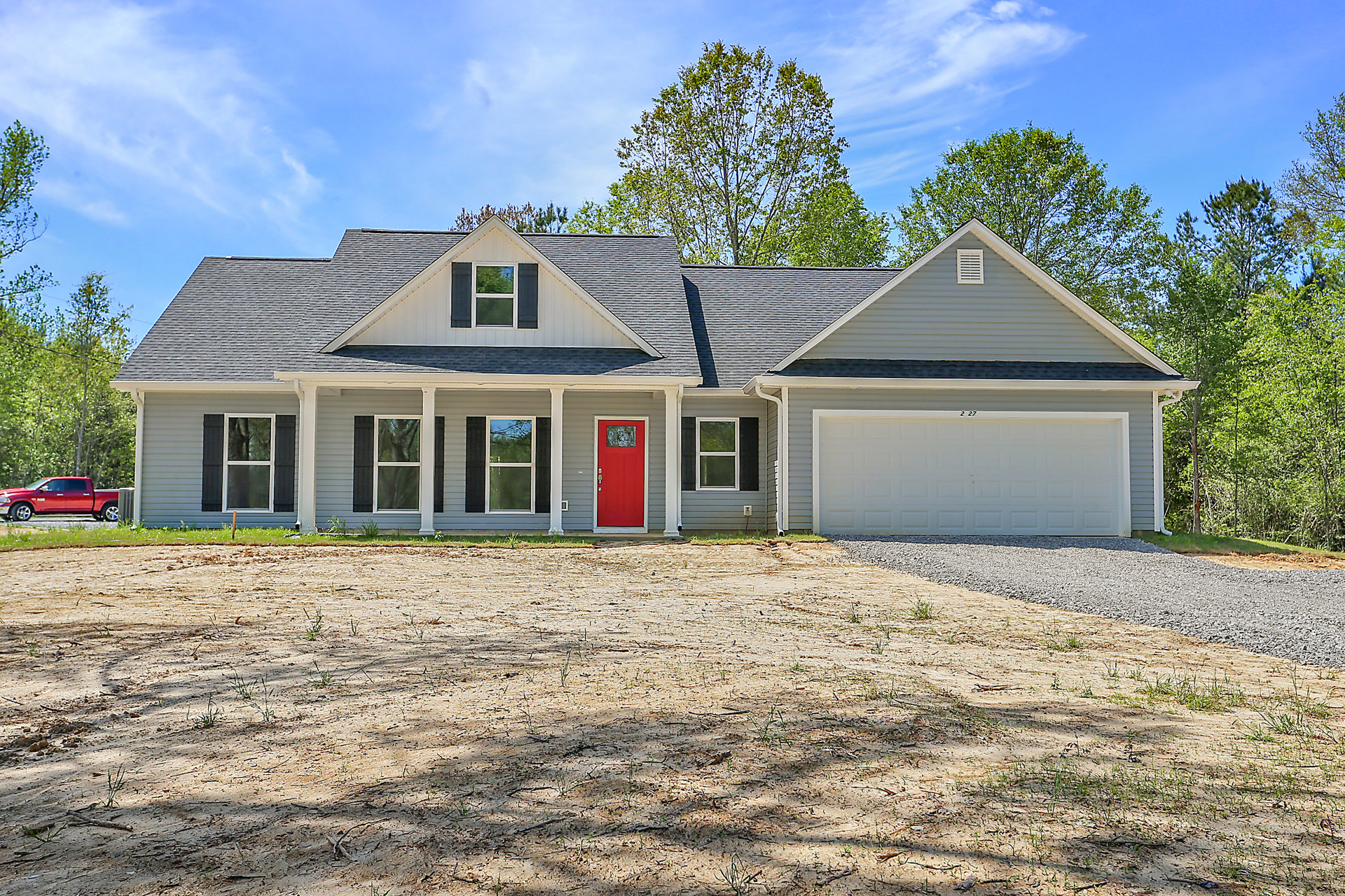 Two-story house with white siding, red front door with glass window, white-framed windows, attached white garage door, red car parked in driveway, tree and dirt field in