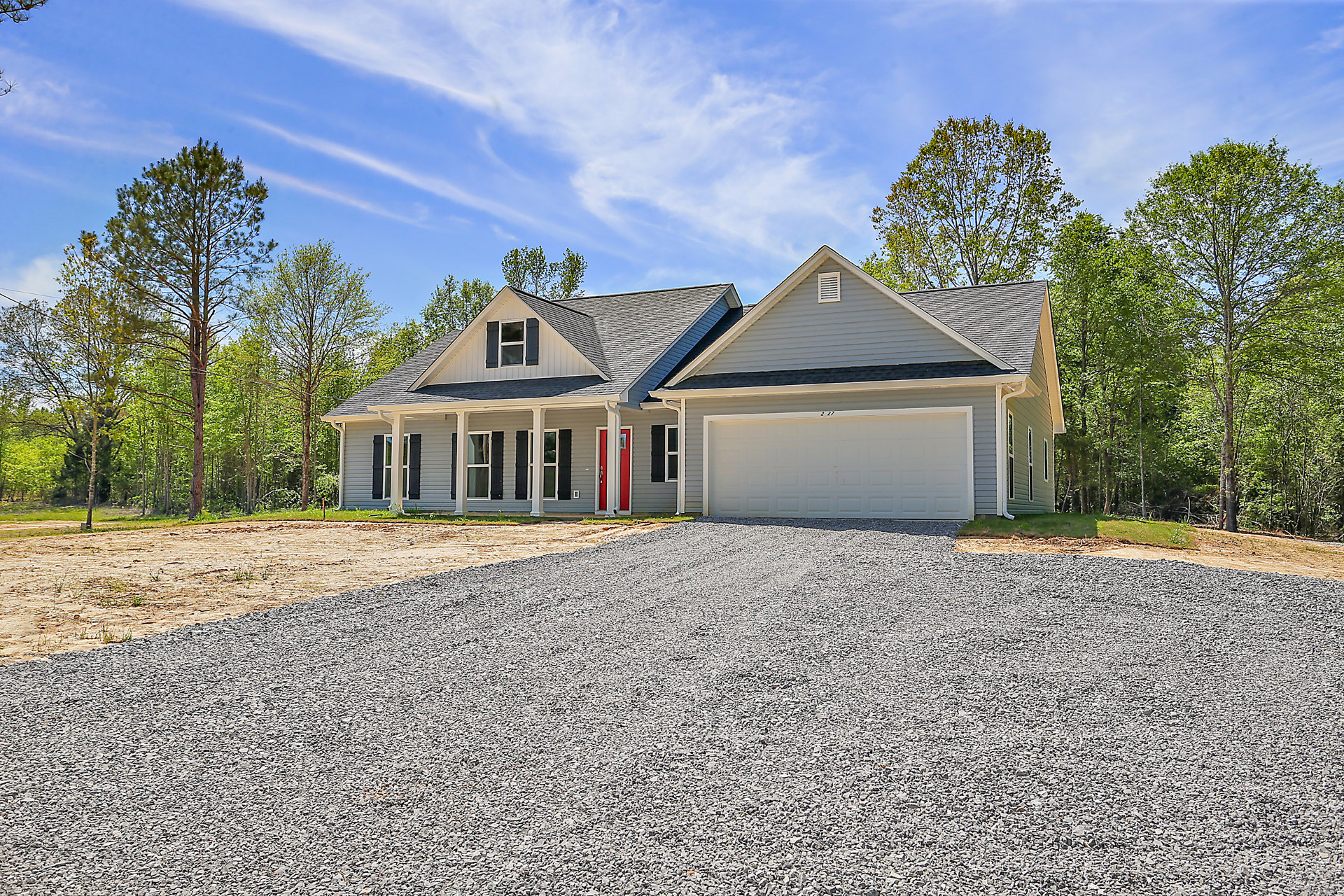Two-story home with white siding, attached garage, gravel driveway, and mature leafy trees in front yard under partly cloudy sky