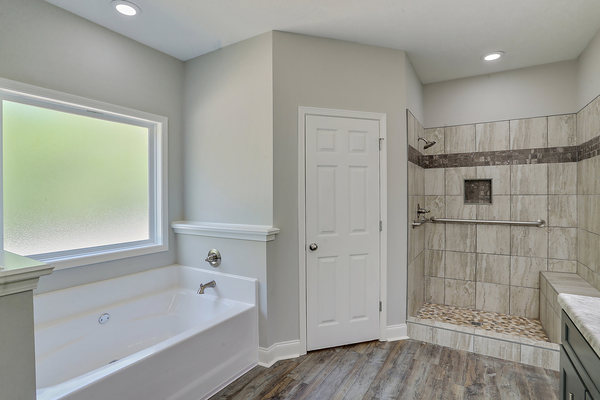 White freestanding bathtub with silver faucets beside glass-enclosed shower, white door with silver doorknob, frosted glass window set in stone tile wall, light gray floor tiles
