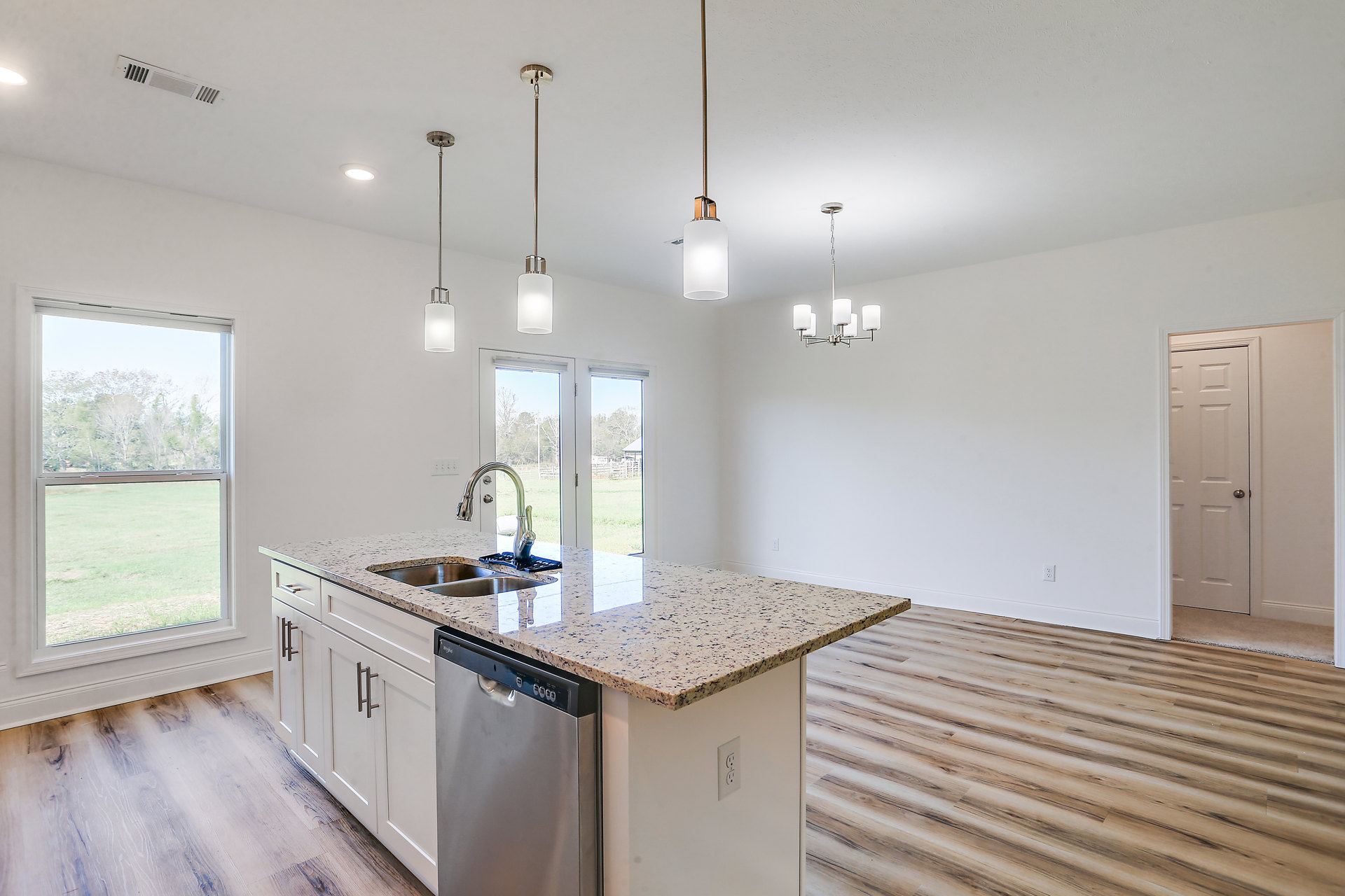 Modern kitchen with white cabinetry, stainless steel sink and dishwasher, stone countertop, window overlooking trees, and white door with silver handle