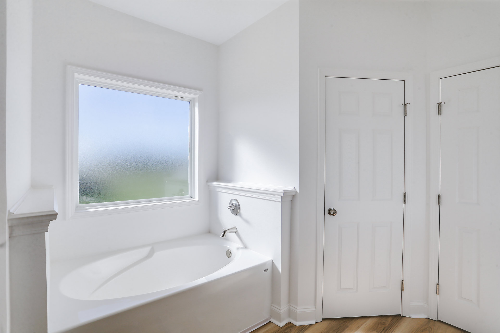 White freestanding bathtub beside a white wall, frosted glass window, and white door with silver knob in a modern bathroom.