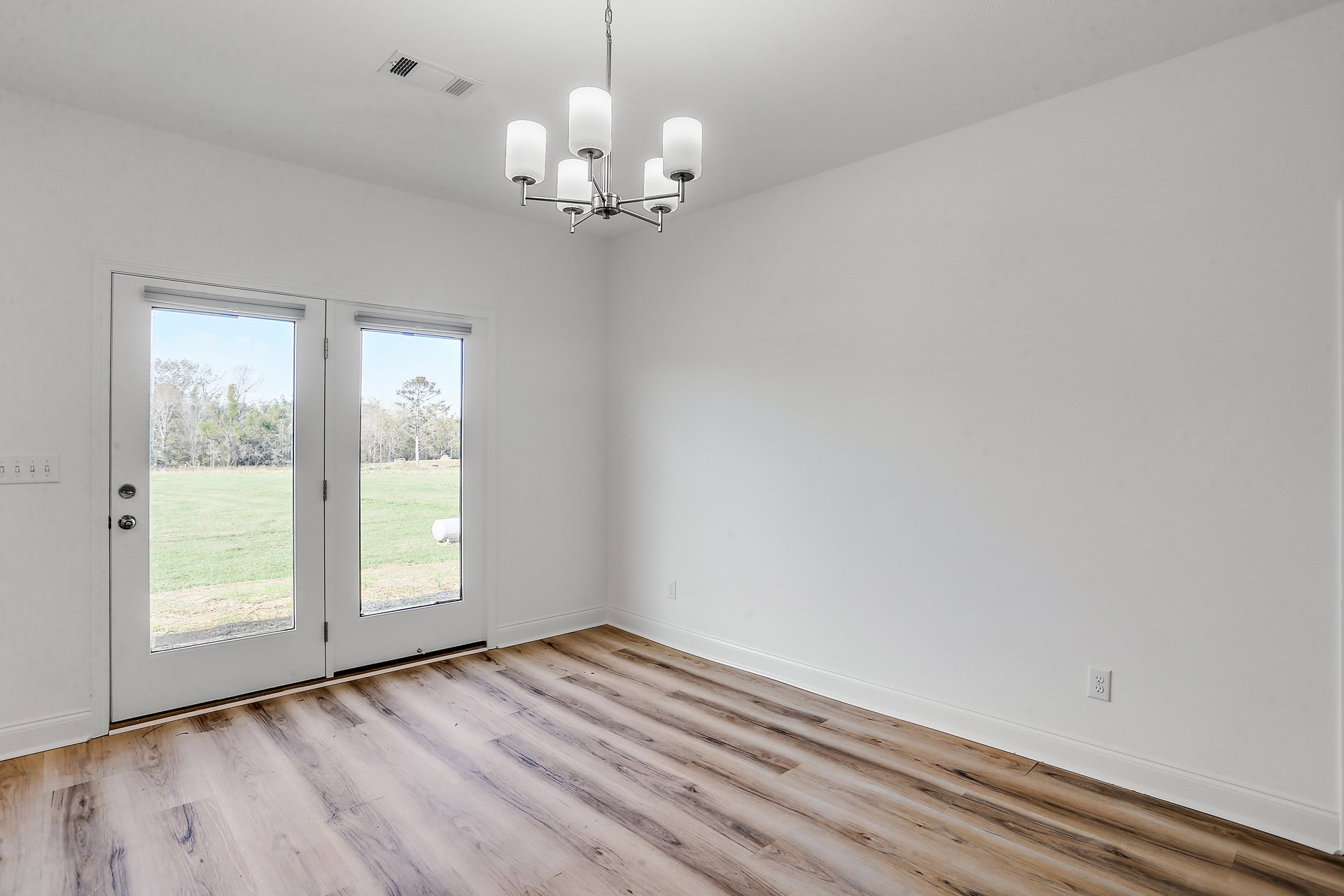 Wood flooring in a spacious room, double glass-paneled doors, white chandelier with silver accents hanging from a plaster ceiling, neutral walls.